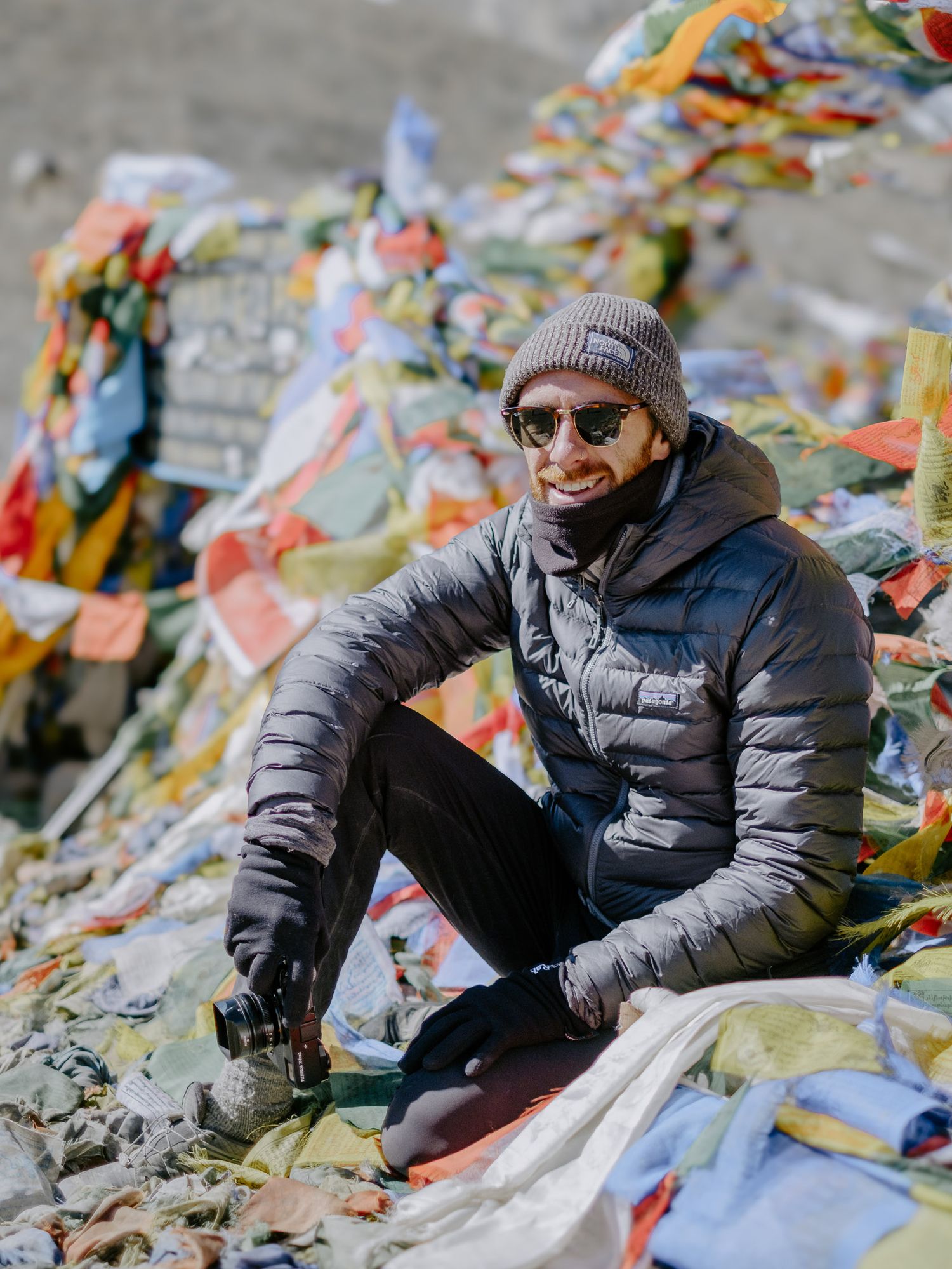 Hiker relaxes among colorful prayer flags at a mountain pass viewpoint.