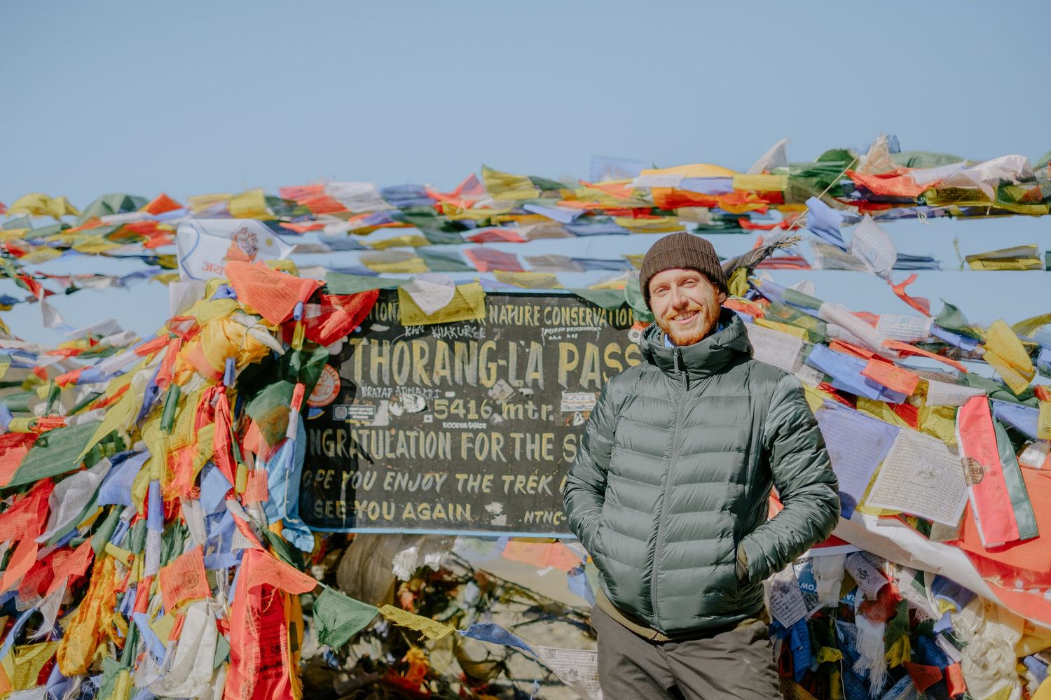 Prayer flags wave near a wooden sign marking Thorong La Pass in the mountains.