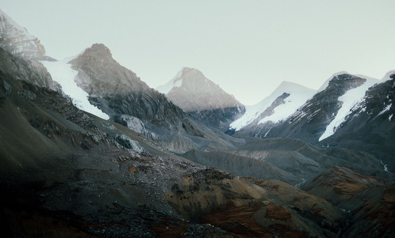 Snow-capped mountain peaks rise dramatically against a moody sky in a rugged mountain landscape.