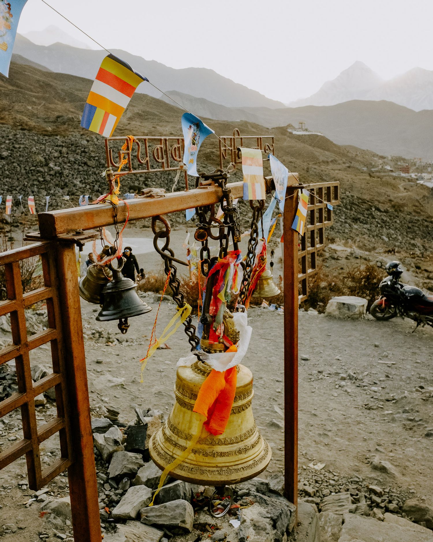 Buddhist prayer flags and bells adorn a mountain summit against a misty mountain landscape in the Himalayas.