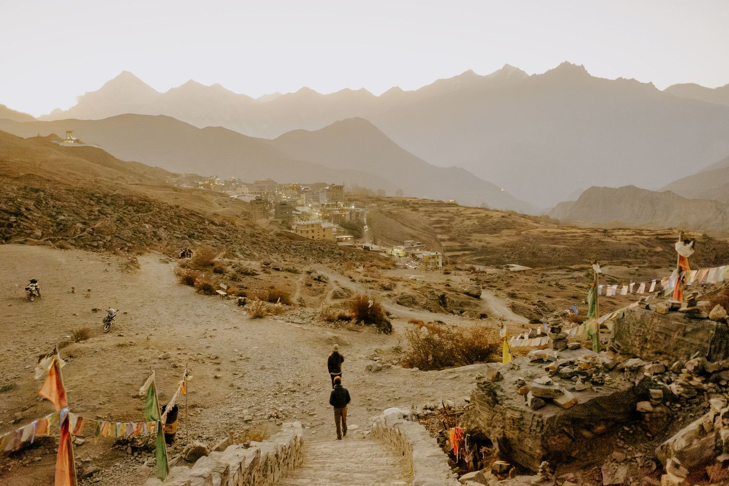 A panoramic view of hikers traversing a rugged mountain trail with distant peaks and desert landscape in the background.