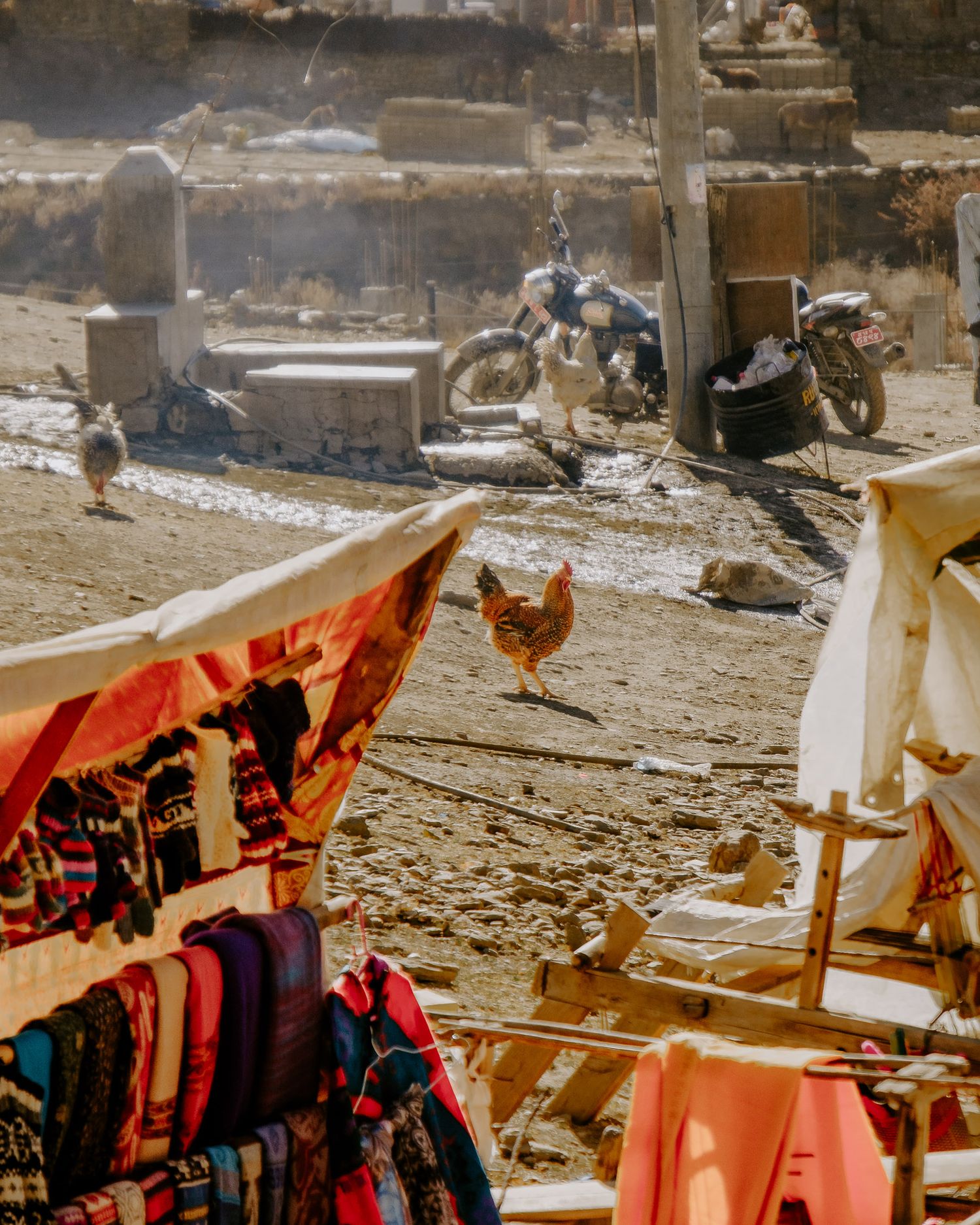 Colorful clothing and textiles hang on display at an outdoor market with chickens roaming nearby.