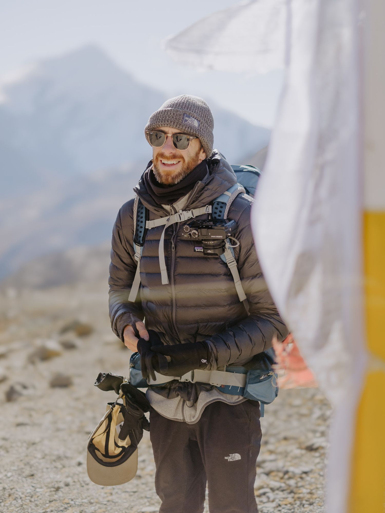 A hiker in winter gear stands on a mountain trail with prayer flags visible.