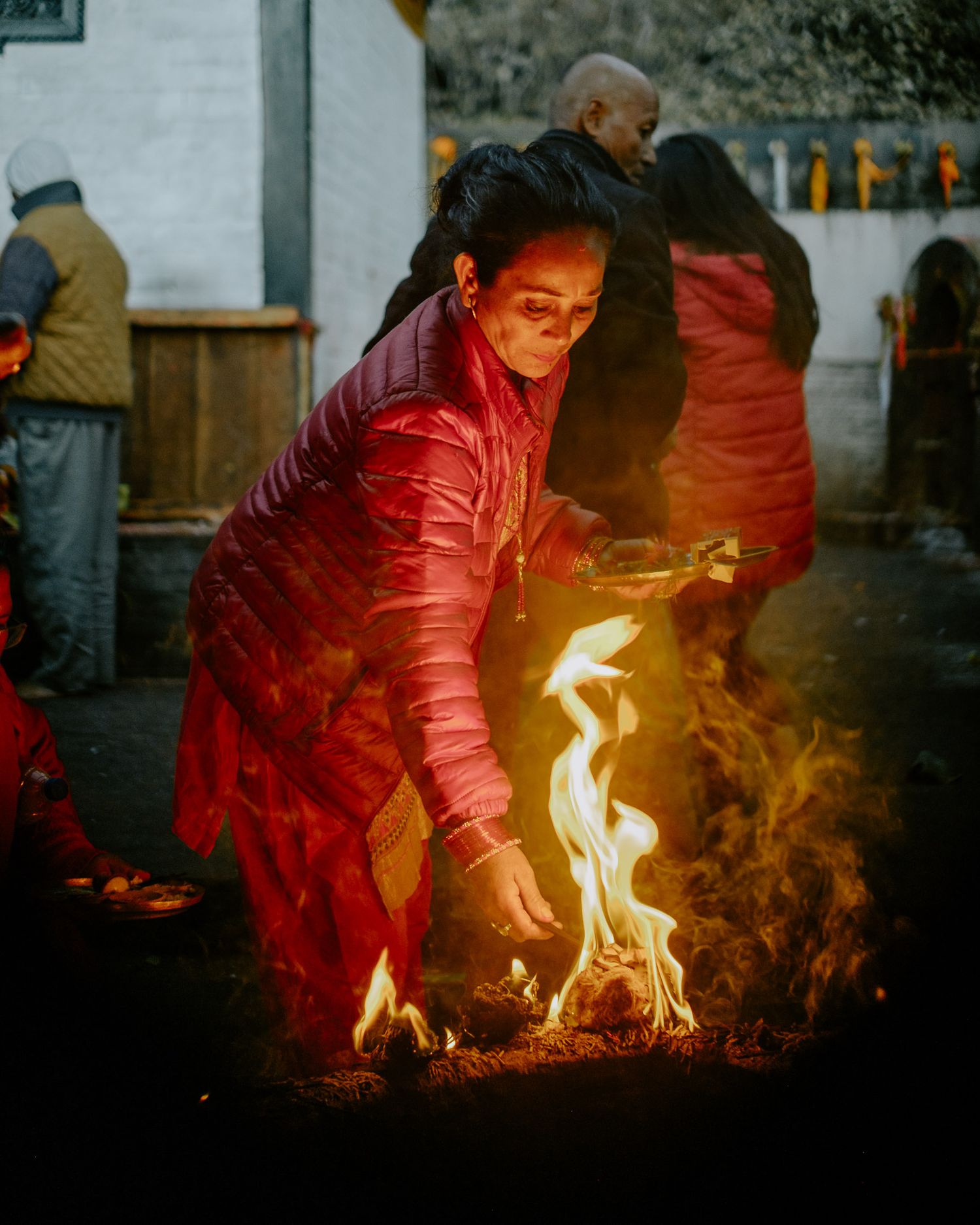 A person in a red jacket tends to glowing flames and embers in a dimly lit ritual or ceremonial setting.