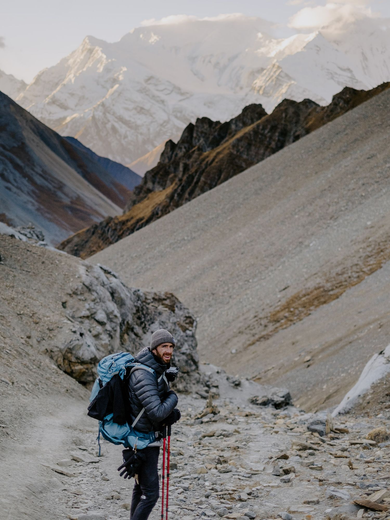 Mountaineer climbing steep rocky terrain with snow-capped peaks visible in the distance.