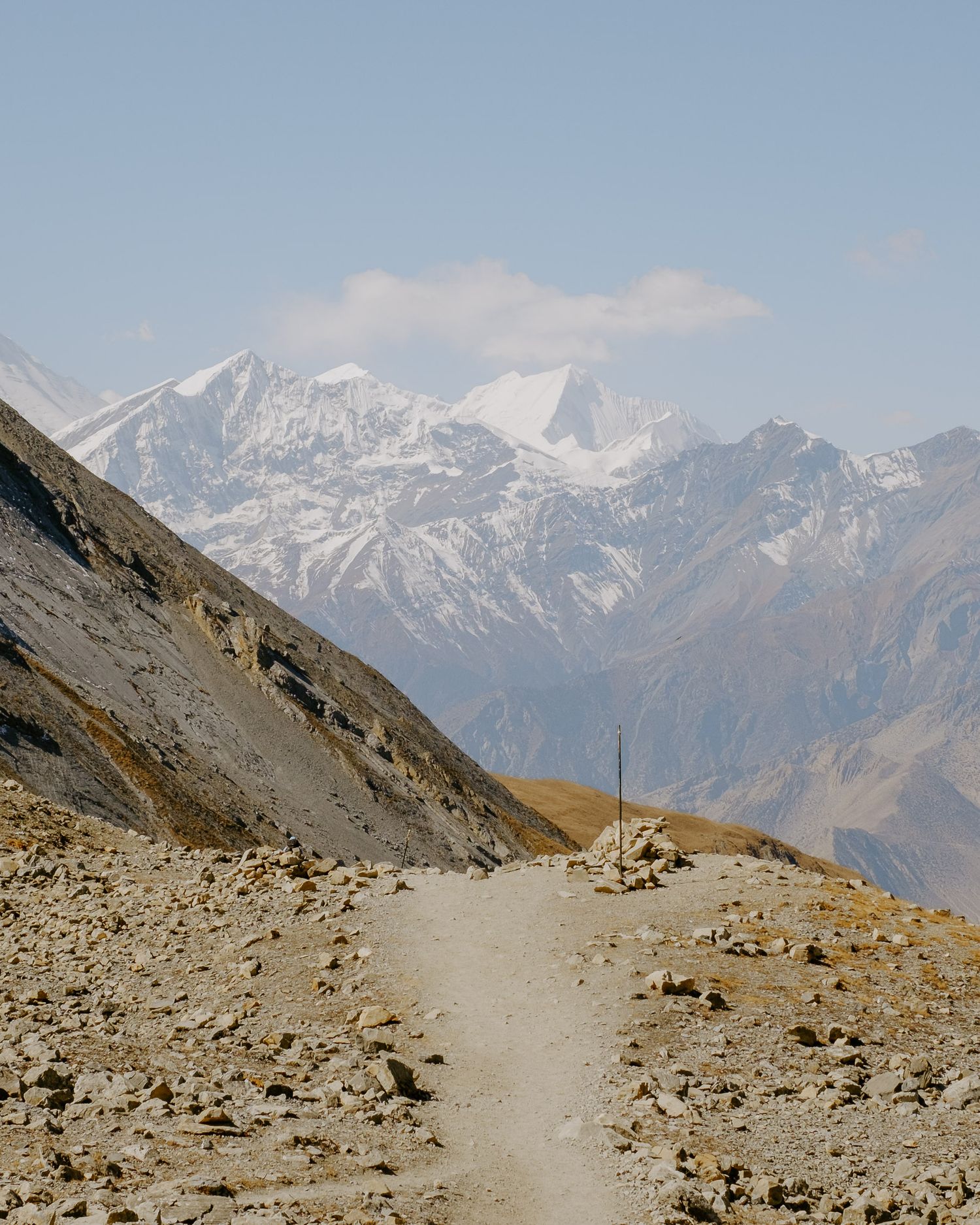 Snow-capped mountain peaks rise above a winding dirt trail through barren high-altitude landscape.
