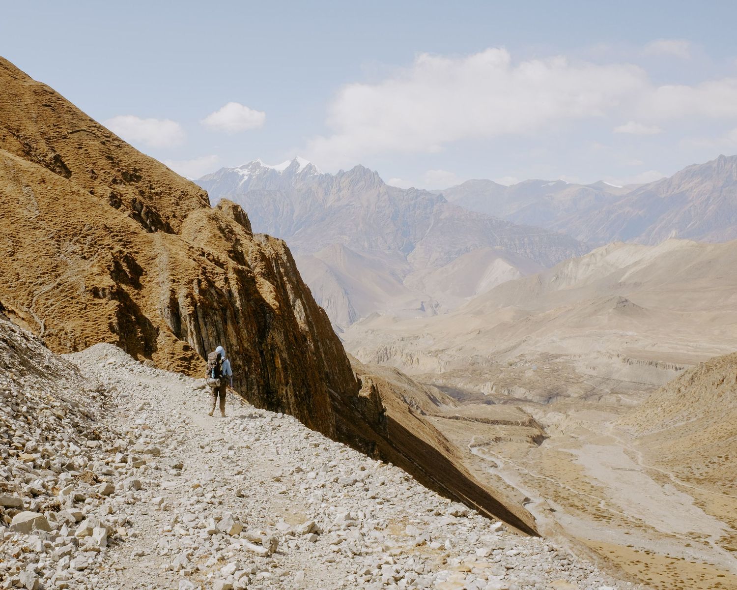 Hikers traverse a rocky mountain trail with dramatic peaks and valleys in the background.