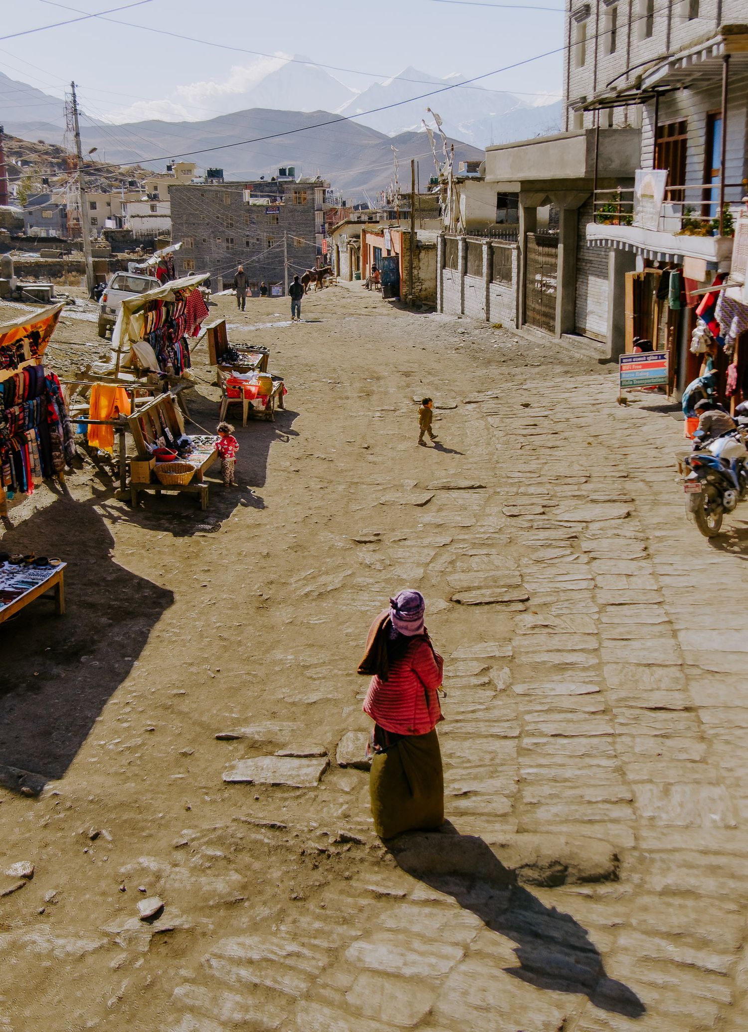A street scene in a mountainous town with vendors and pedestrians walking along a stone-paved road.