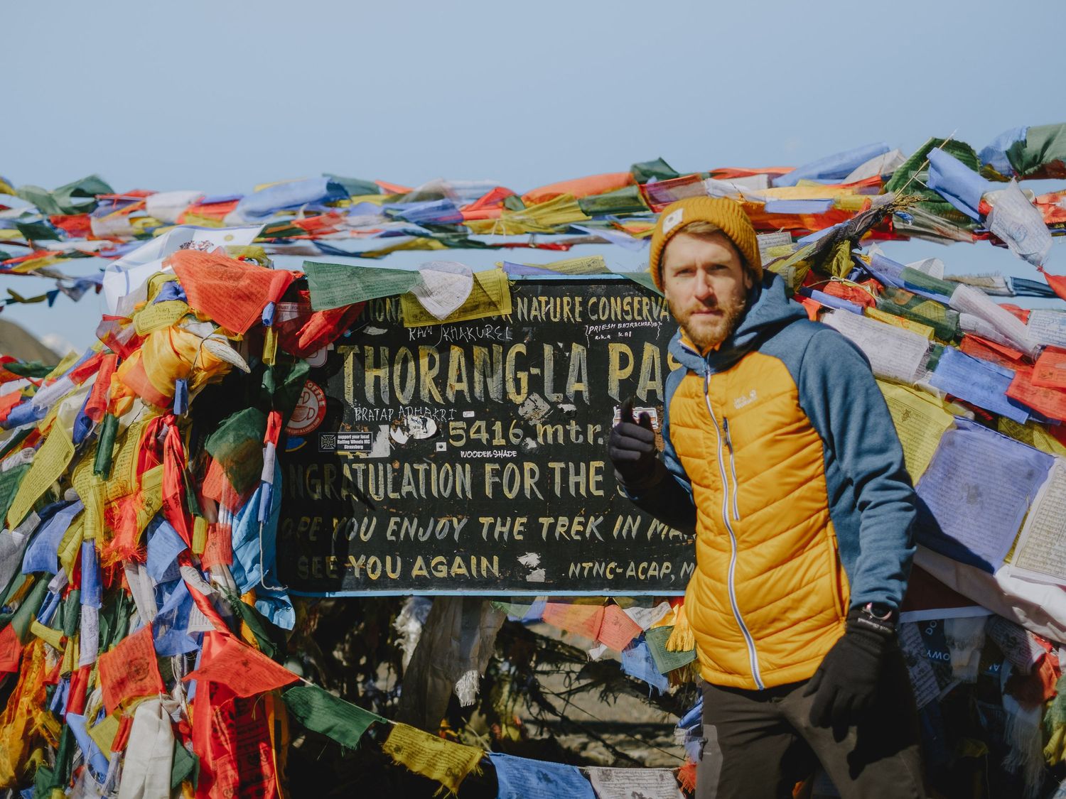 Hikers stand near a wooden trail marker surrounded by colorful prayer flags in the mountains.
