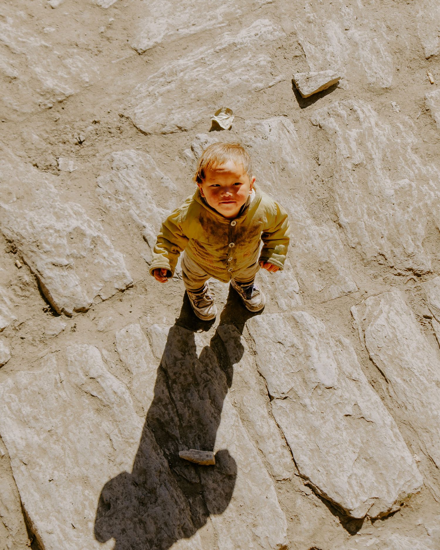 A figure crouches on light-colored rocky ground casting a strong shadow in natural sunlight.