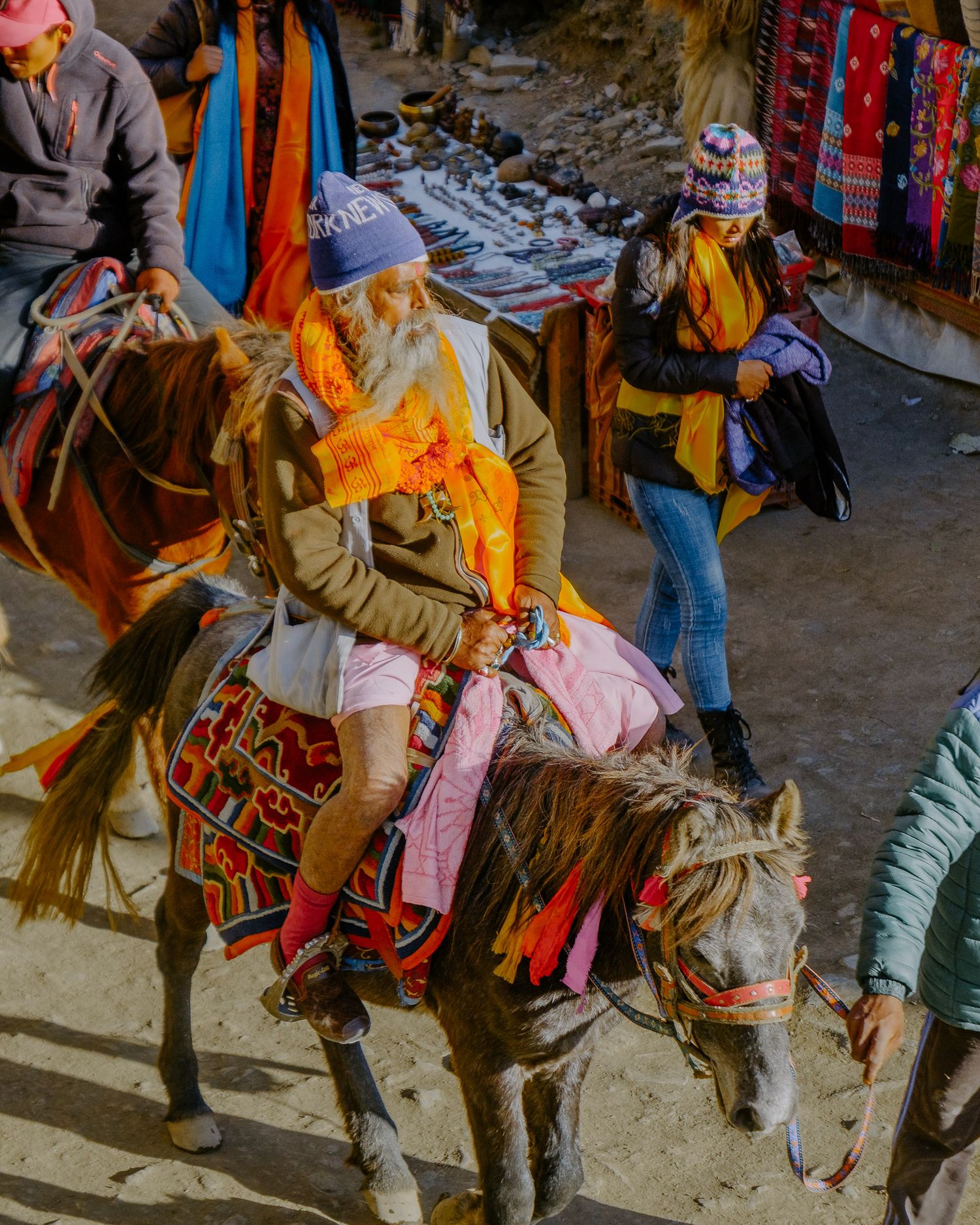 Decorated horses with colorful saddles and tassels stand in a line at a mountain marketplace.