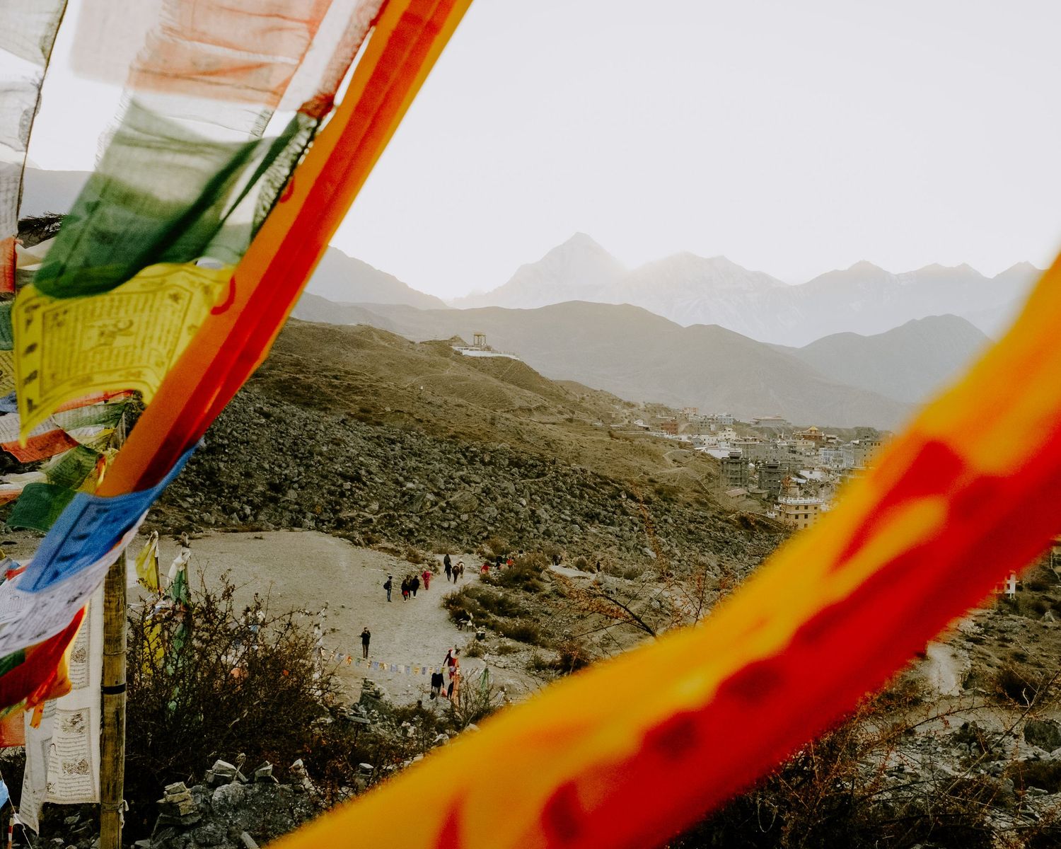 Colorful prayer flags frame distant mountain landscape with hikers on winding path below.