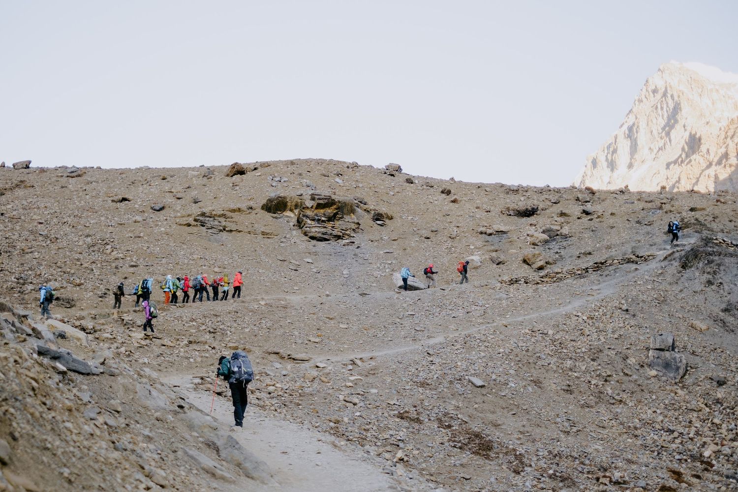 Hikers traversing a barren mountain slope with dramatic rocky peaks in the background.