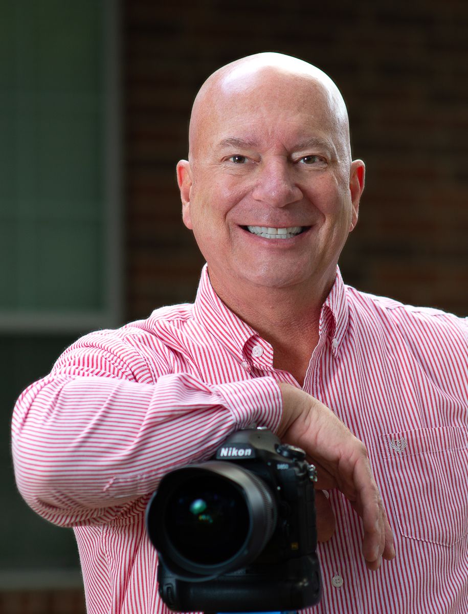 Smiling bald man in pink striped shirt holding a Nikon DSLR camera, posing outdoors as a professional photographer.
