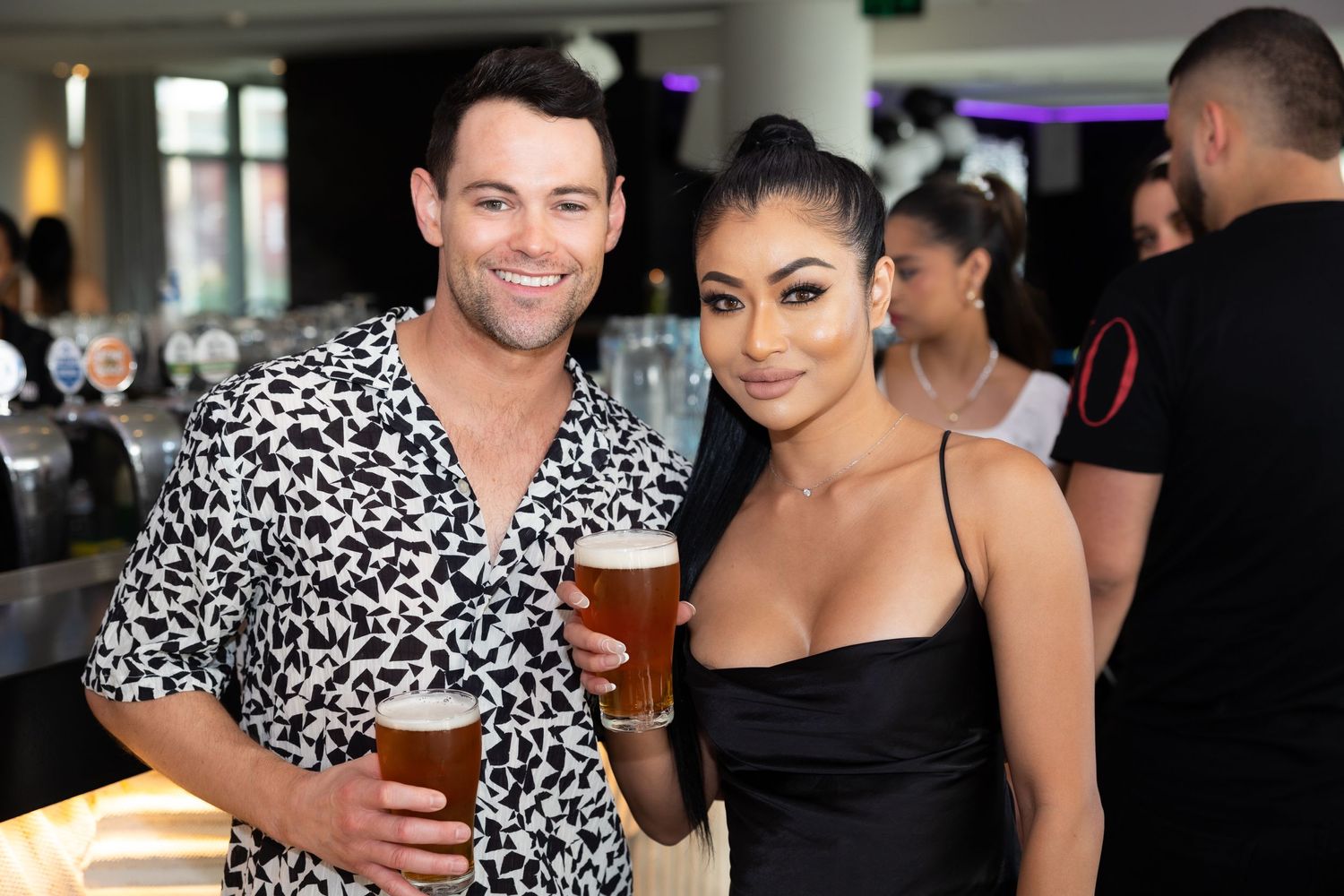 Two people enjoying drinks together at a social gathering, with one wearing a black and white patterned shirt.
