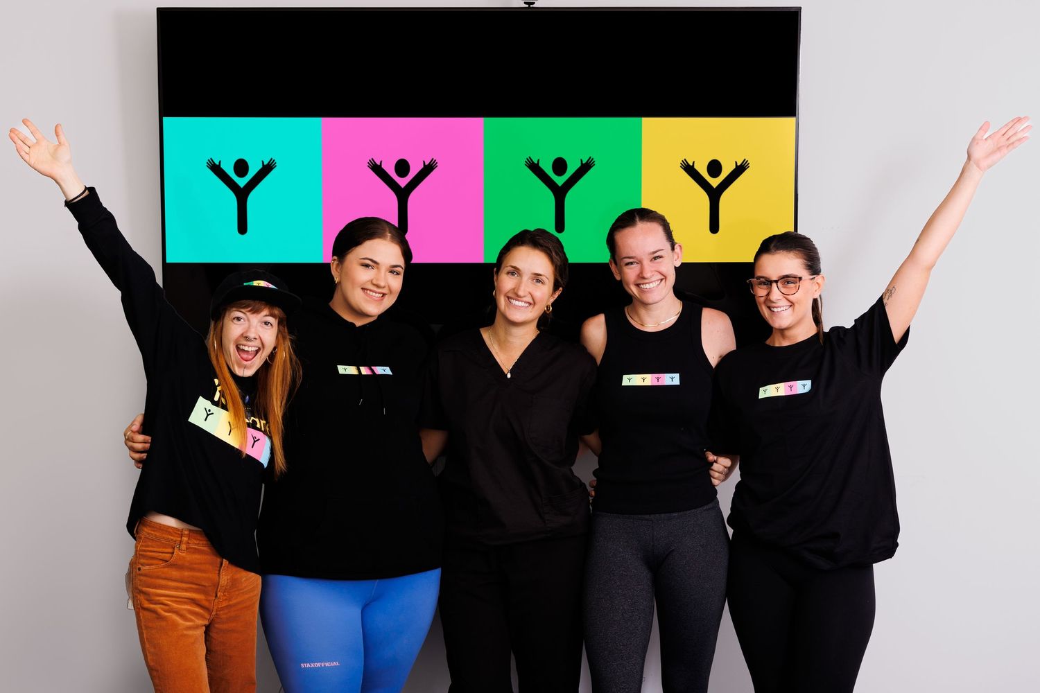 Team members in matching dark outfits celebrate with raised arms in front of a vibrant logo wall.