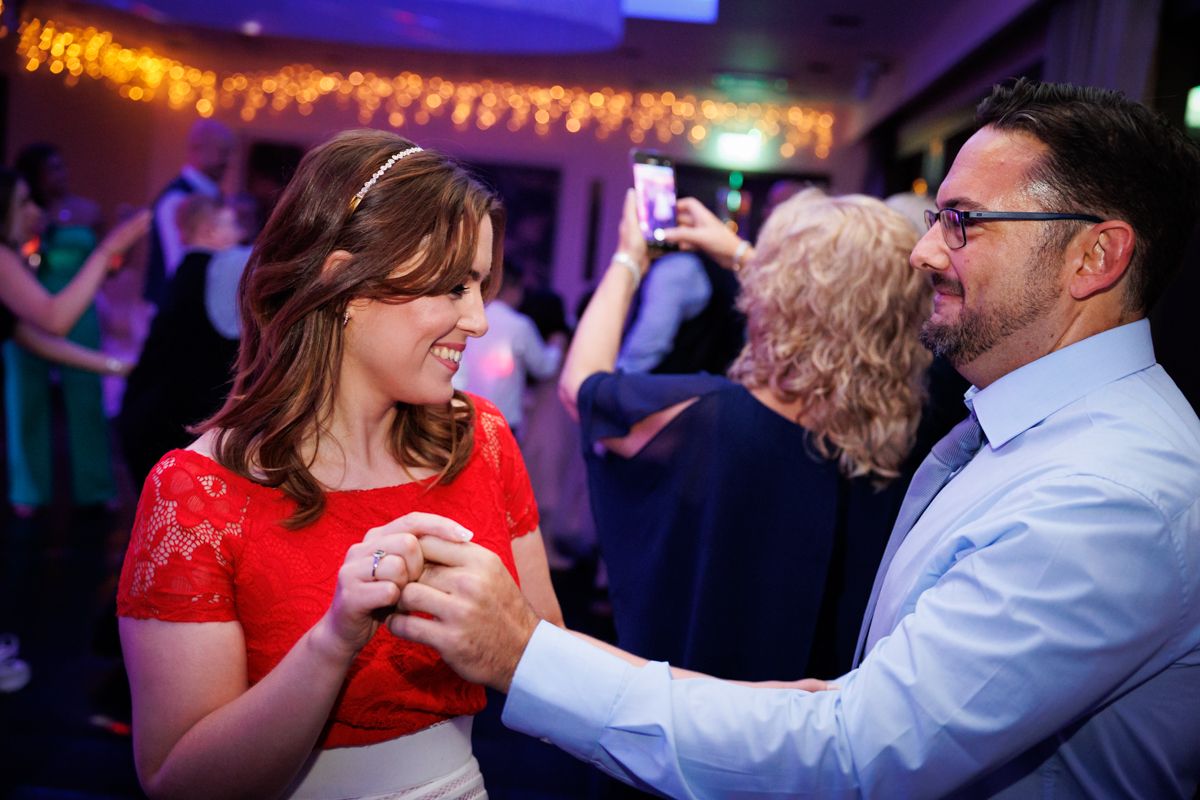 A couple dances together at an evening party under string lights, with the woman wearing a bright red dress.