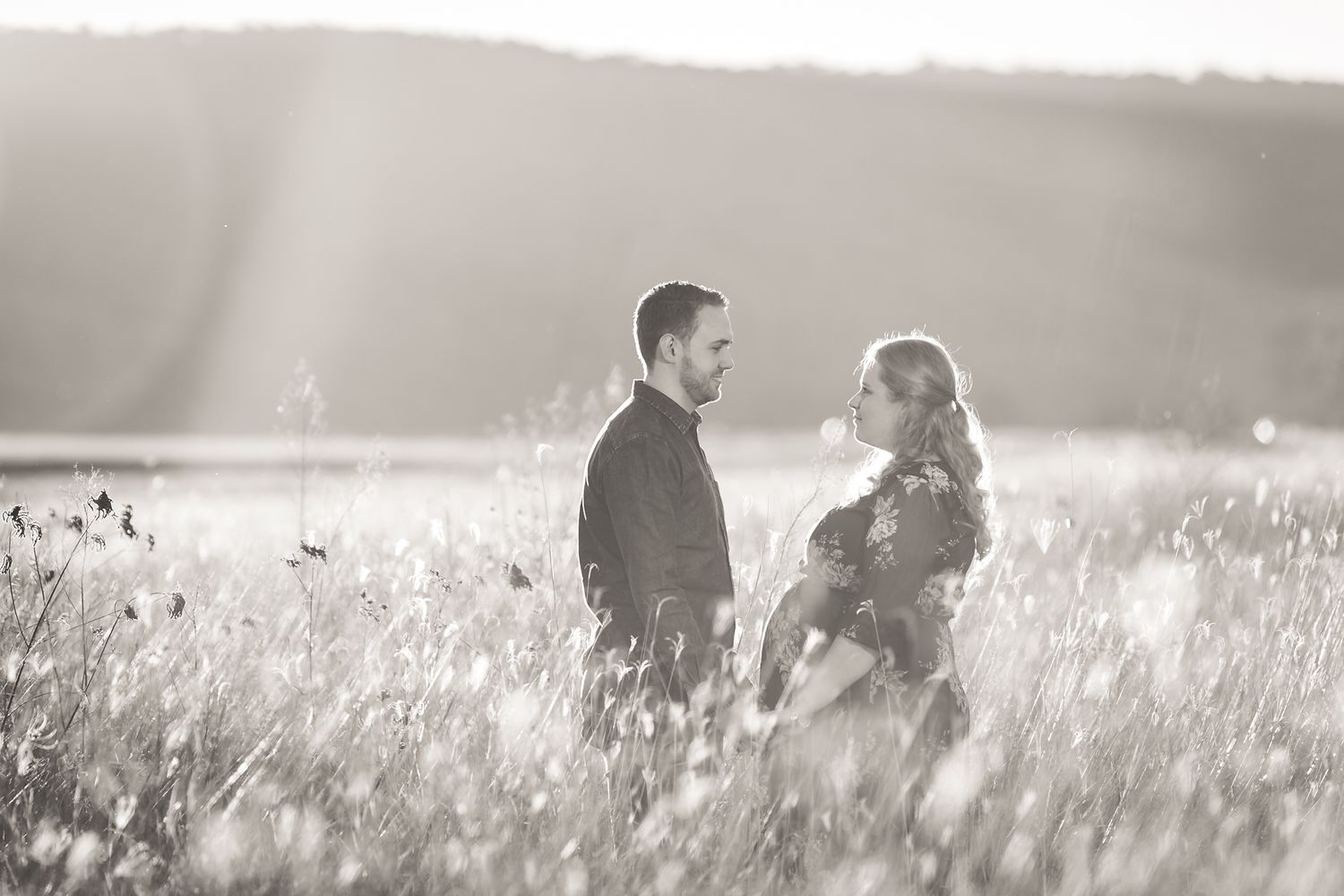 A couple stands together in a sunlit field of tall grass during golden hour, creating a romantic silhouette.