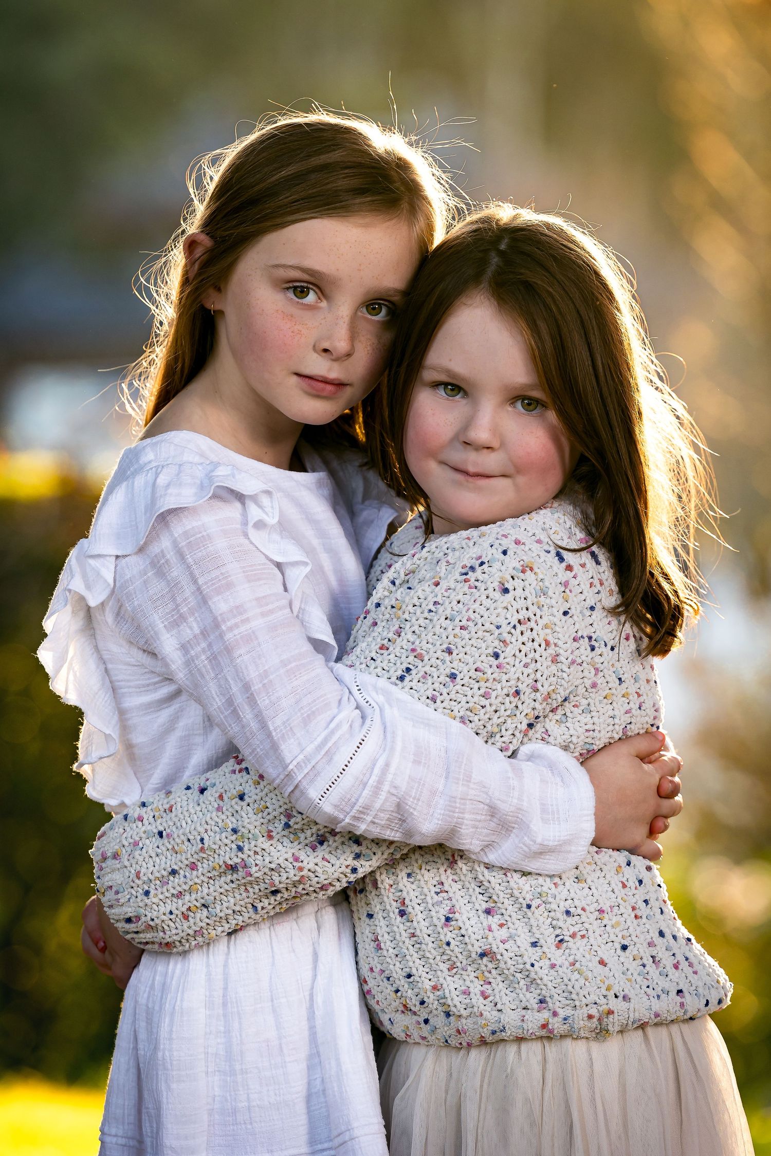 Two children embrace outdoors in natural backlit lighting wearing white dress and knit sweater with dreamy golden backdrop.