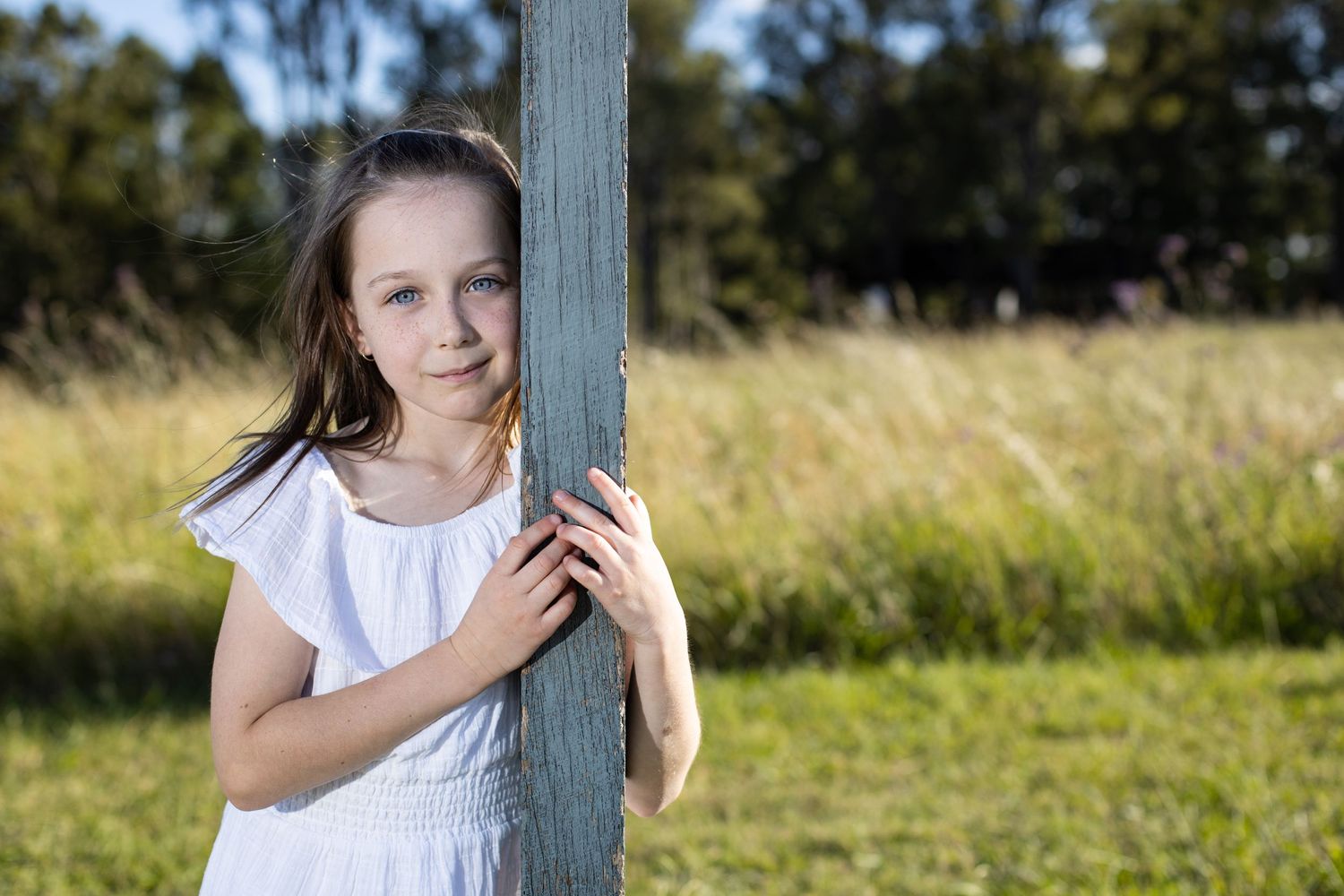 Young person in a white shirt leans against a wooden post in a sunny field with tall grass in the background.