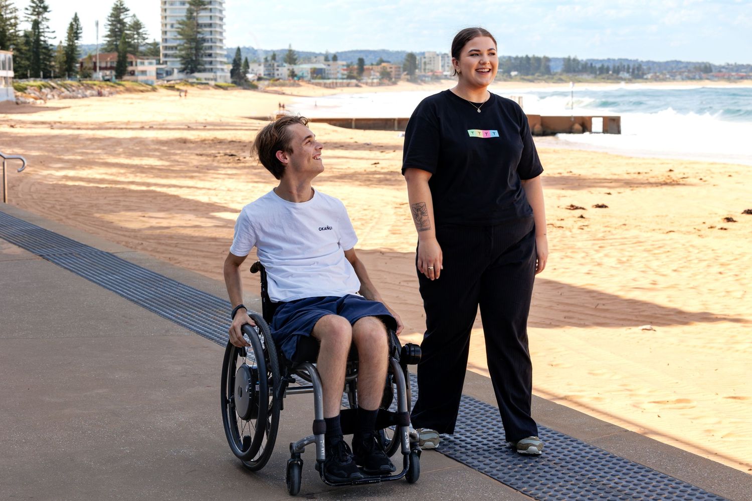 Two people enjoying a stroll along a beachfront boardwalk on a sunny day.