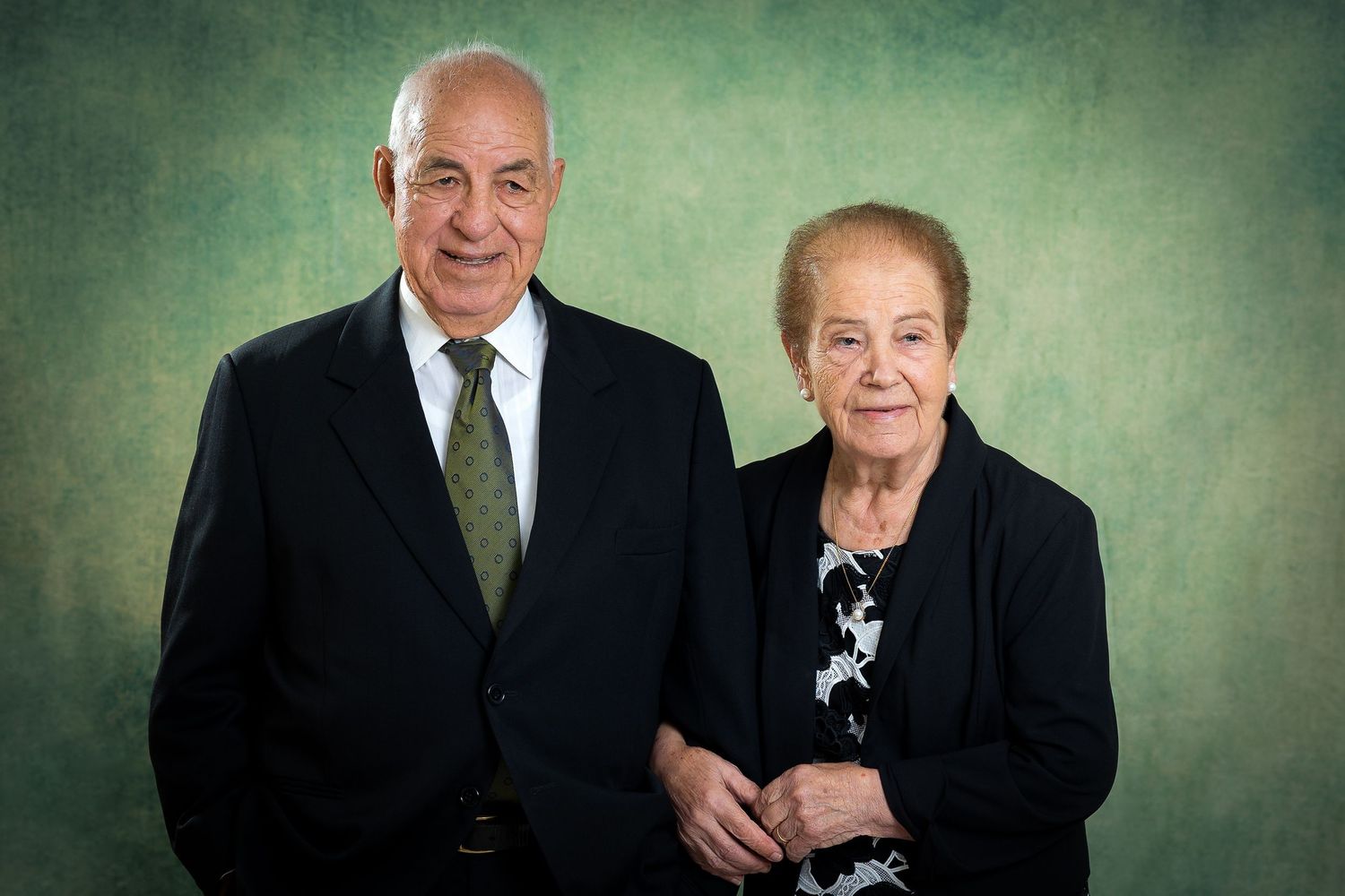 An elderly couple wearing formal attire pose together for a portrait against a textured green background.