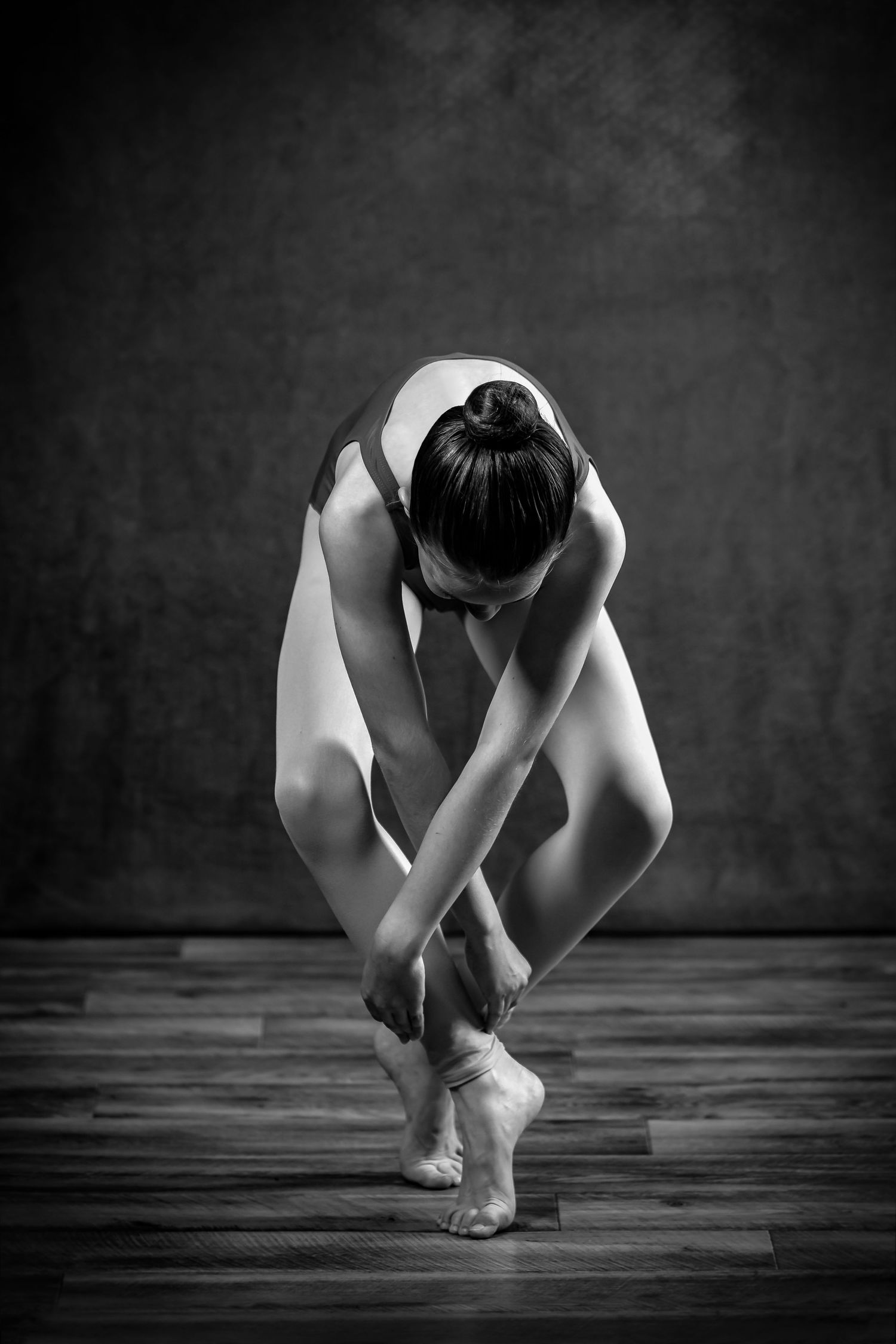 Black and white photo of ballet dancer in contemplative pose on wooden floor.