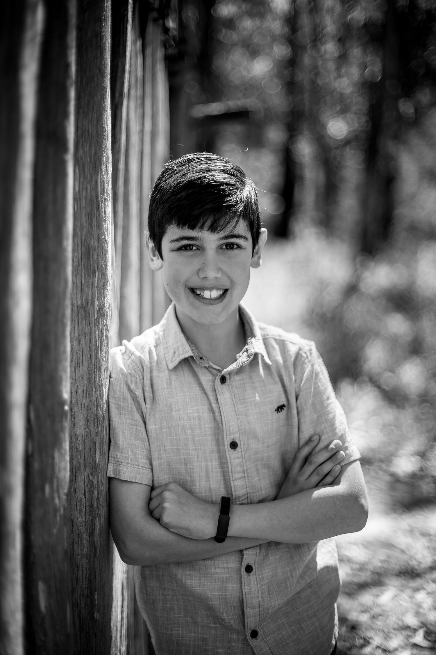Black and white portrait of a young person in a button-down shirt leaning against a wooden post outdoors with blurred trees behind.
