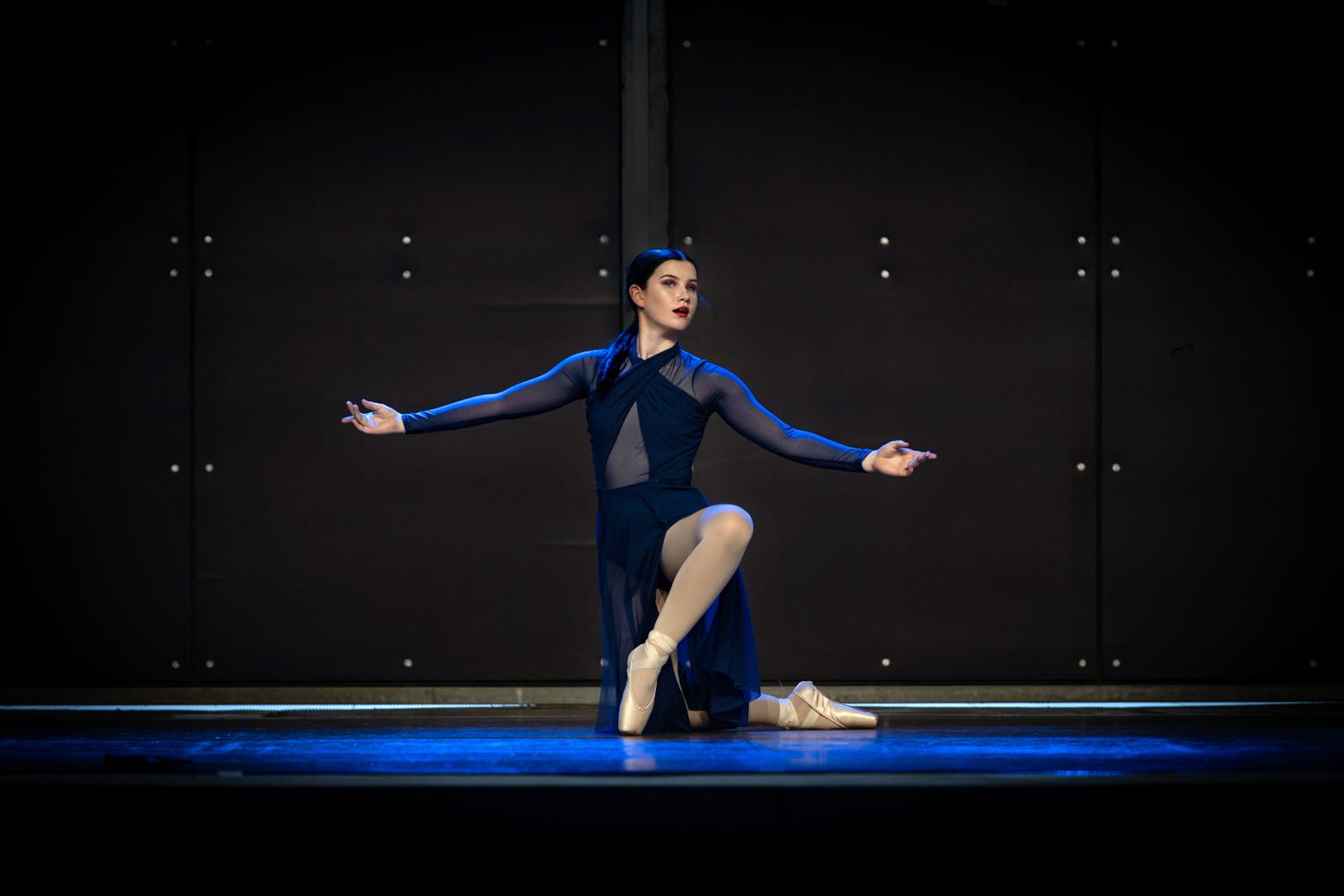 Solo dancer in navy blue costume performing graceful pose on illuminated stage.