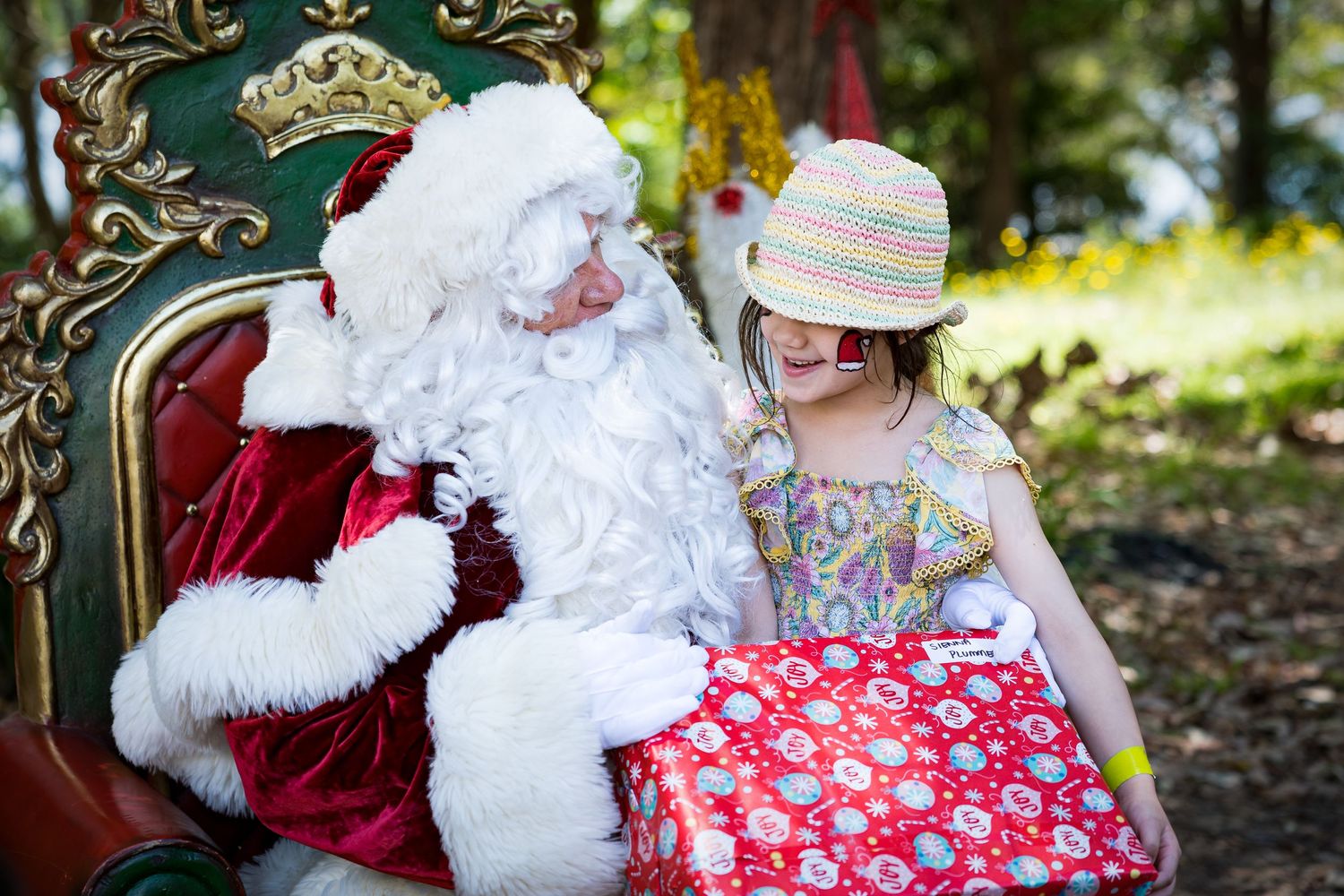 A young child in a summer hat receiving a large red and white wrapped present during an outdoor holiday event.