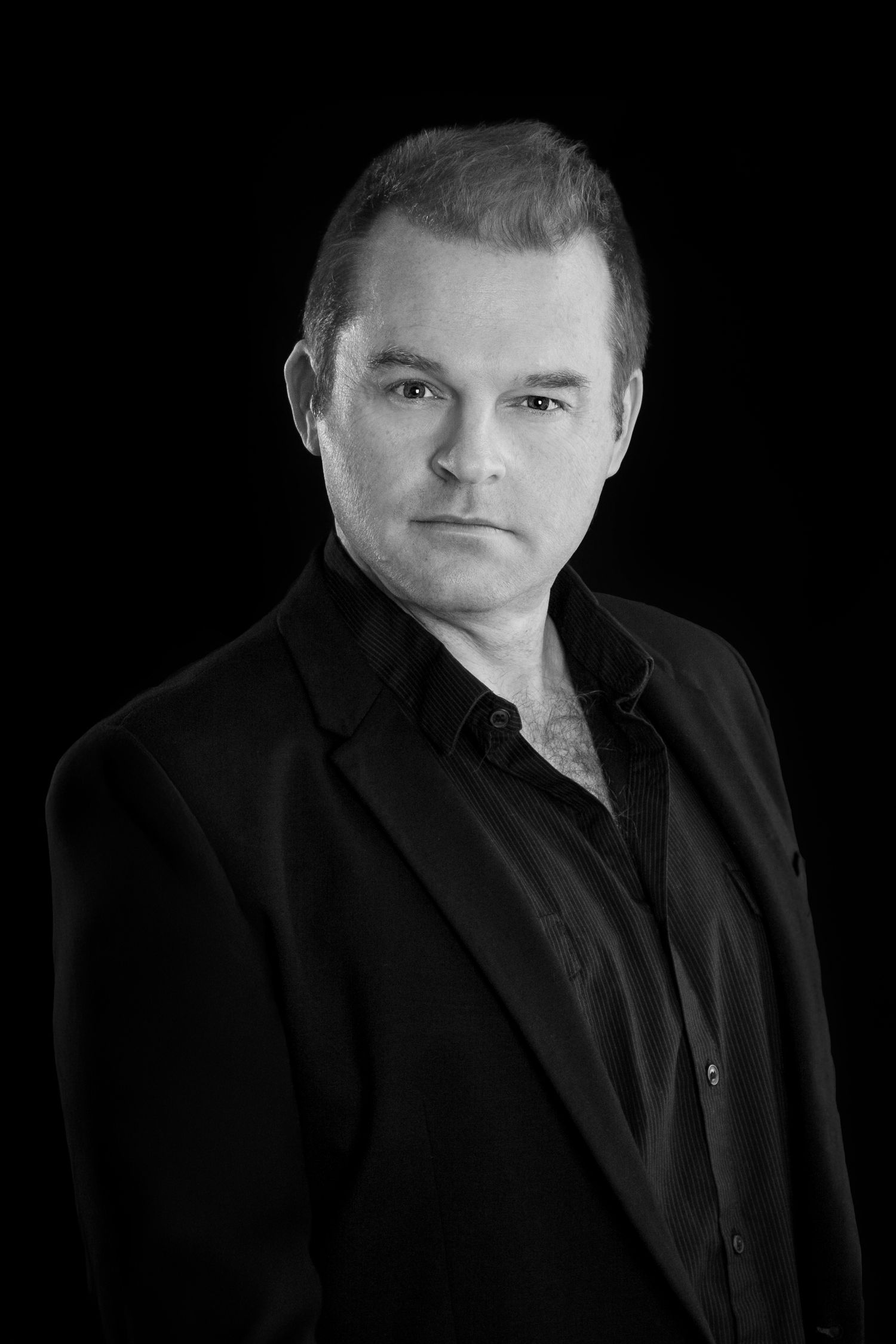 Black and white studio portrait of a person in dark formal attire against black background.