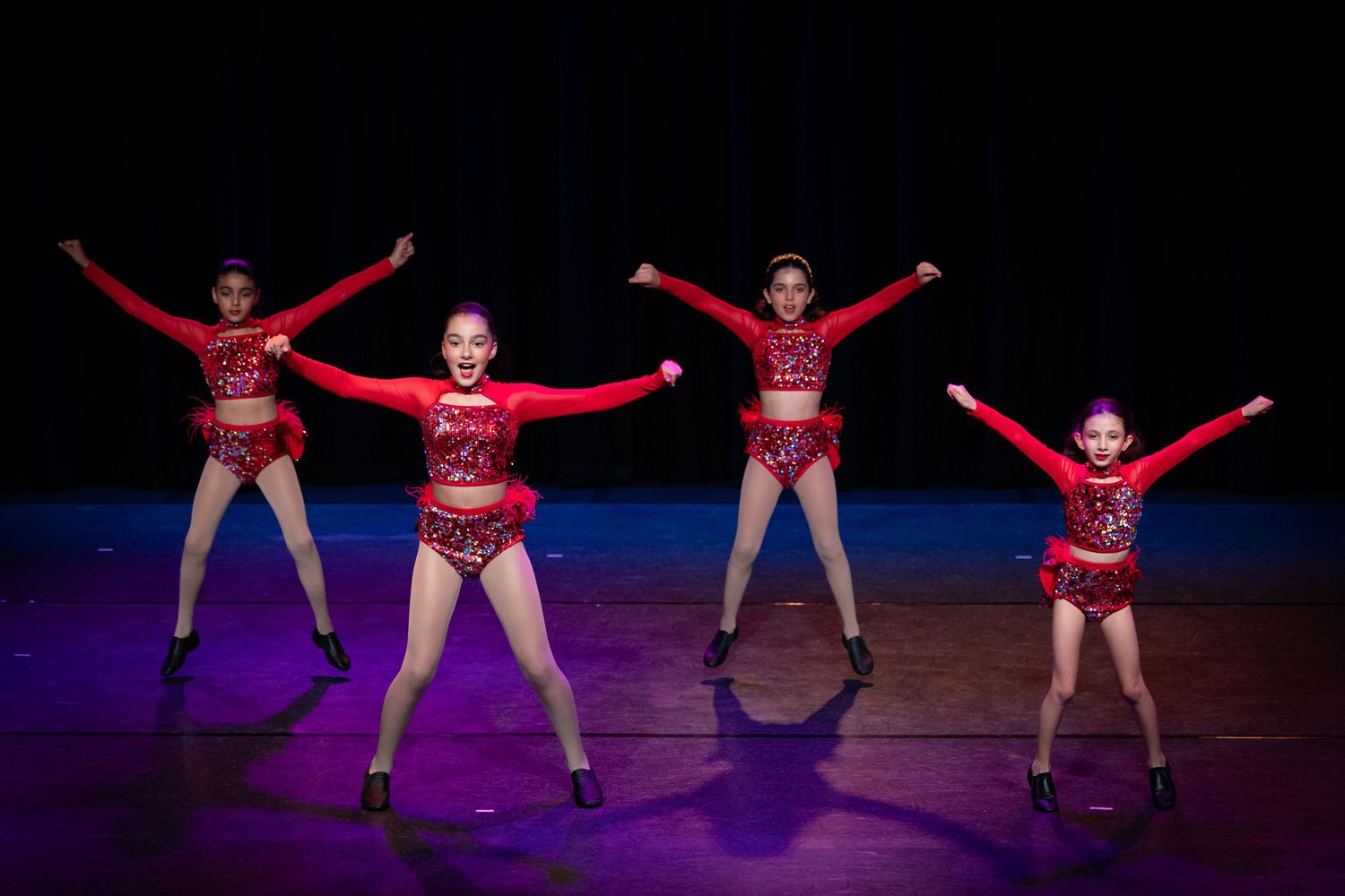 Four dancers in red leotards performing synchronized movements on dark stage.