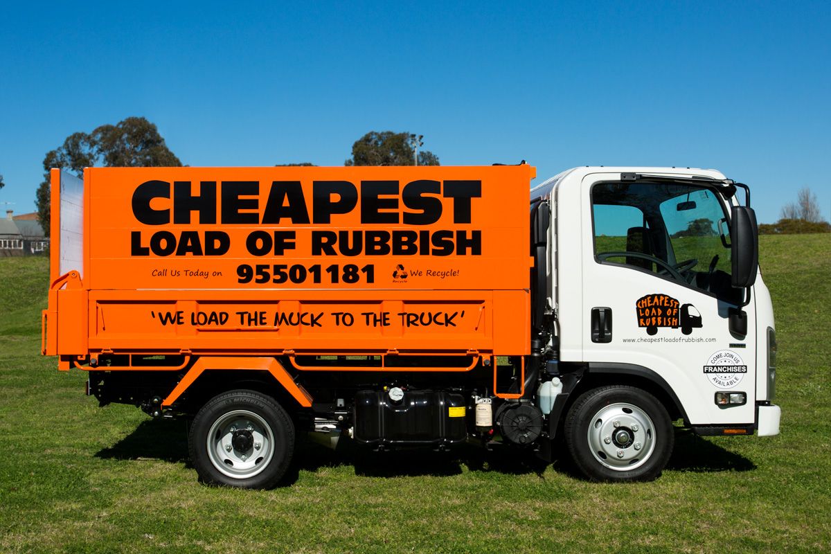 Orange garbage truck with 'Cheapest Load of Rubbish' text parked on grass field.