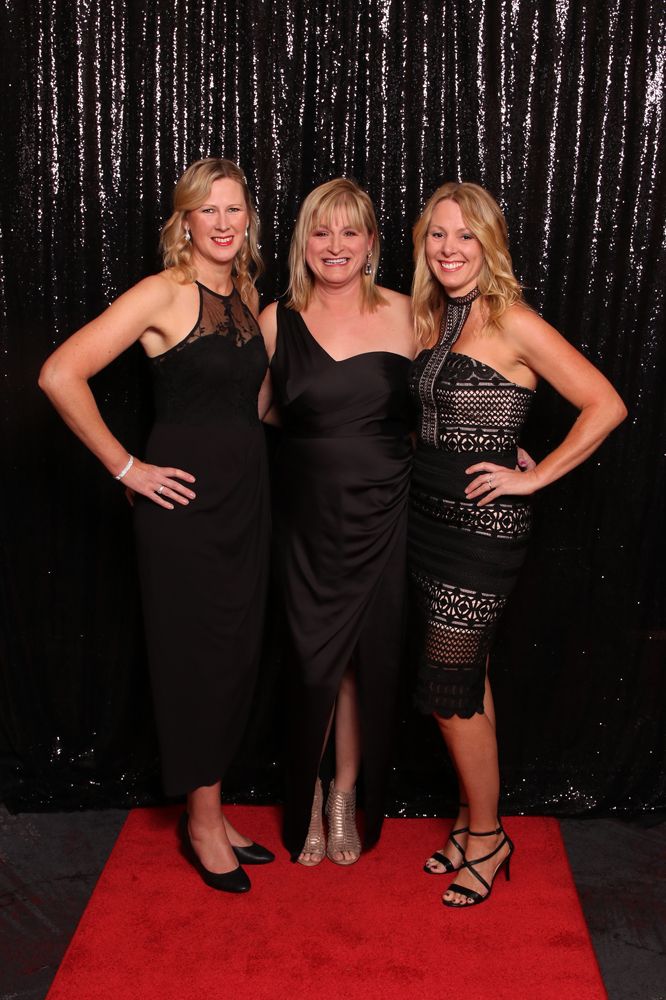 Three women in elegant black dresses pose on red carpet against sequined backdrop.