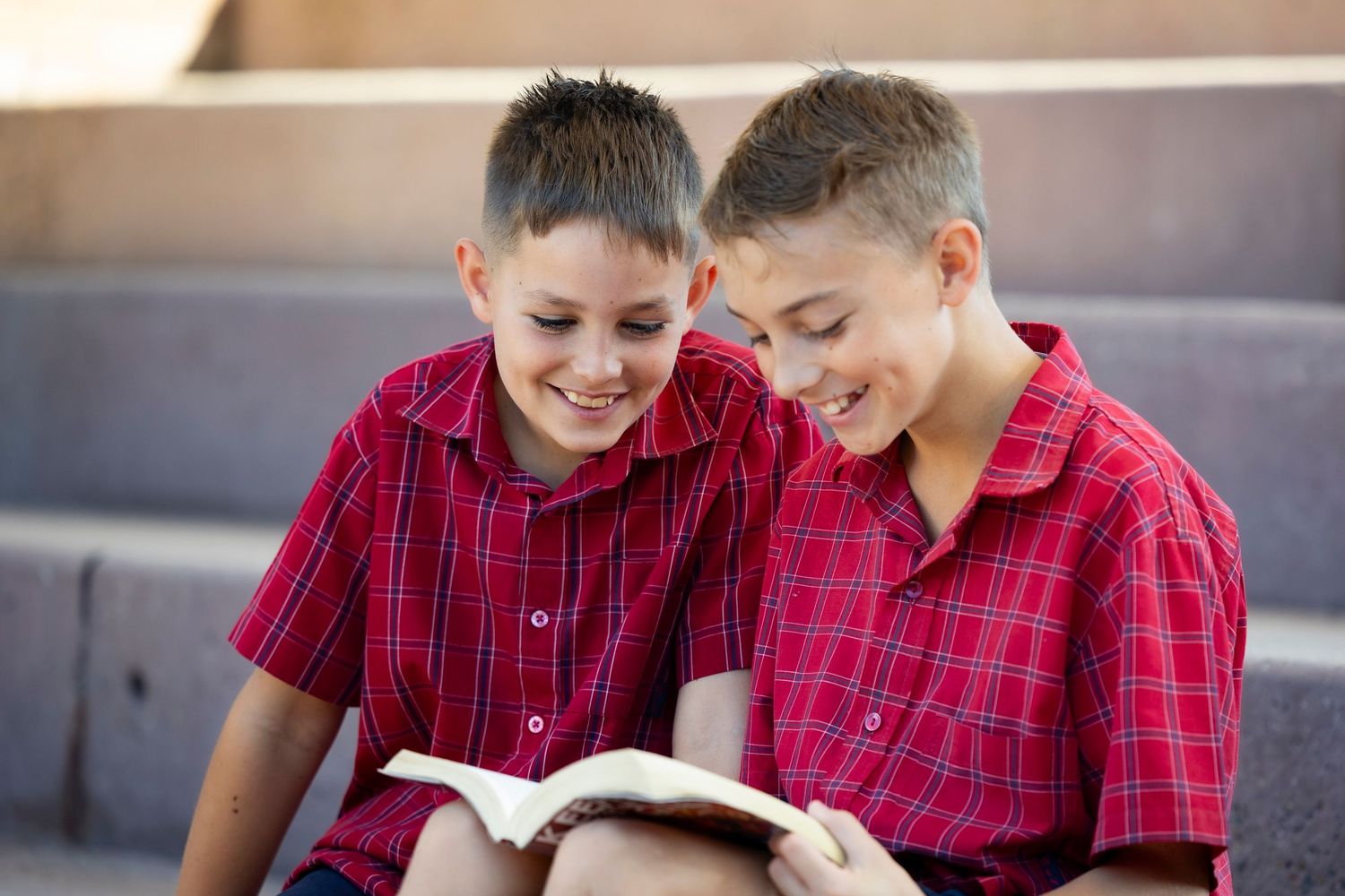 Two students in red plaid uniforms share a book together while sitting on gray steps.