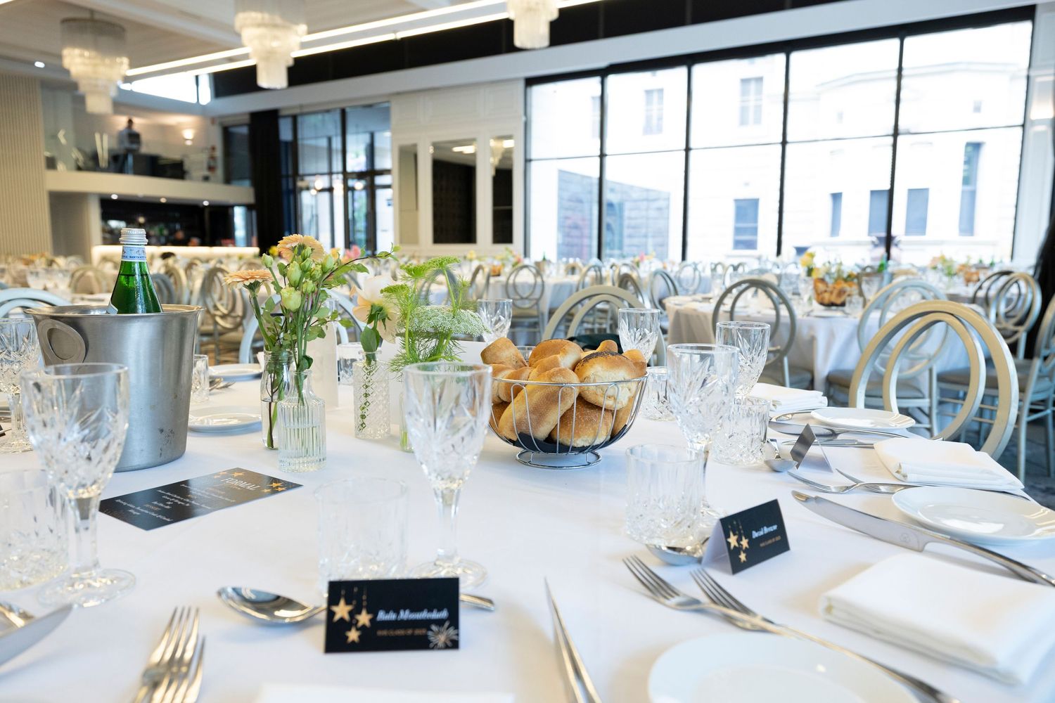 Elegant white table settings with black place cards arranged for a formal dining event in a bright modern restaurant space.