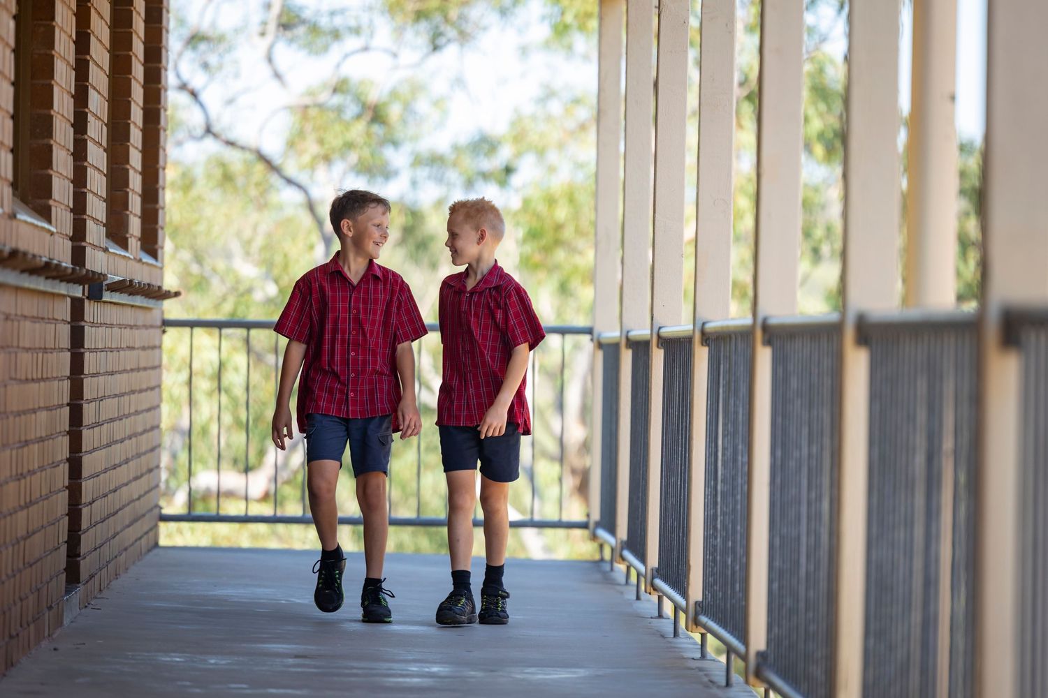 Two students in matching red polo uniforms walk together down a covered outdoor school hallway.