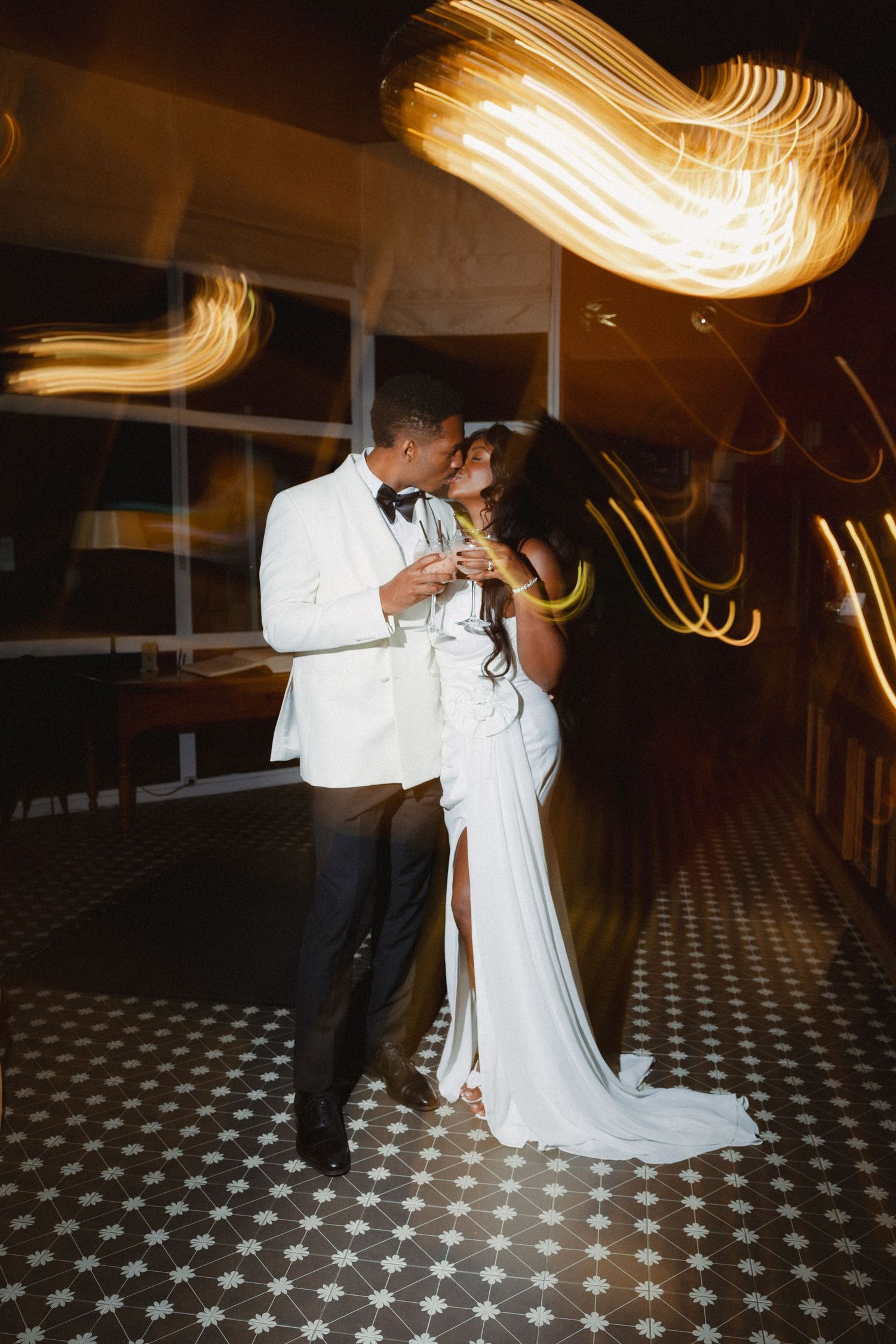 A couple in white formal attire share an intimate dance moment with swirling light trails above on a patterned floor.