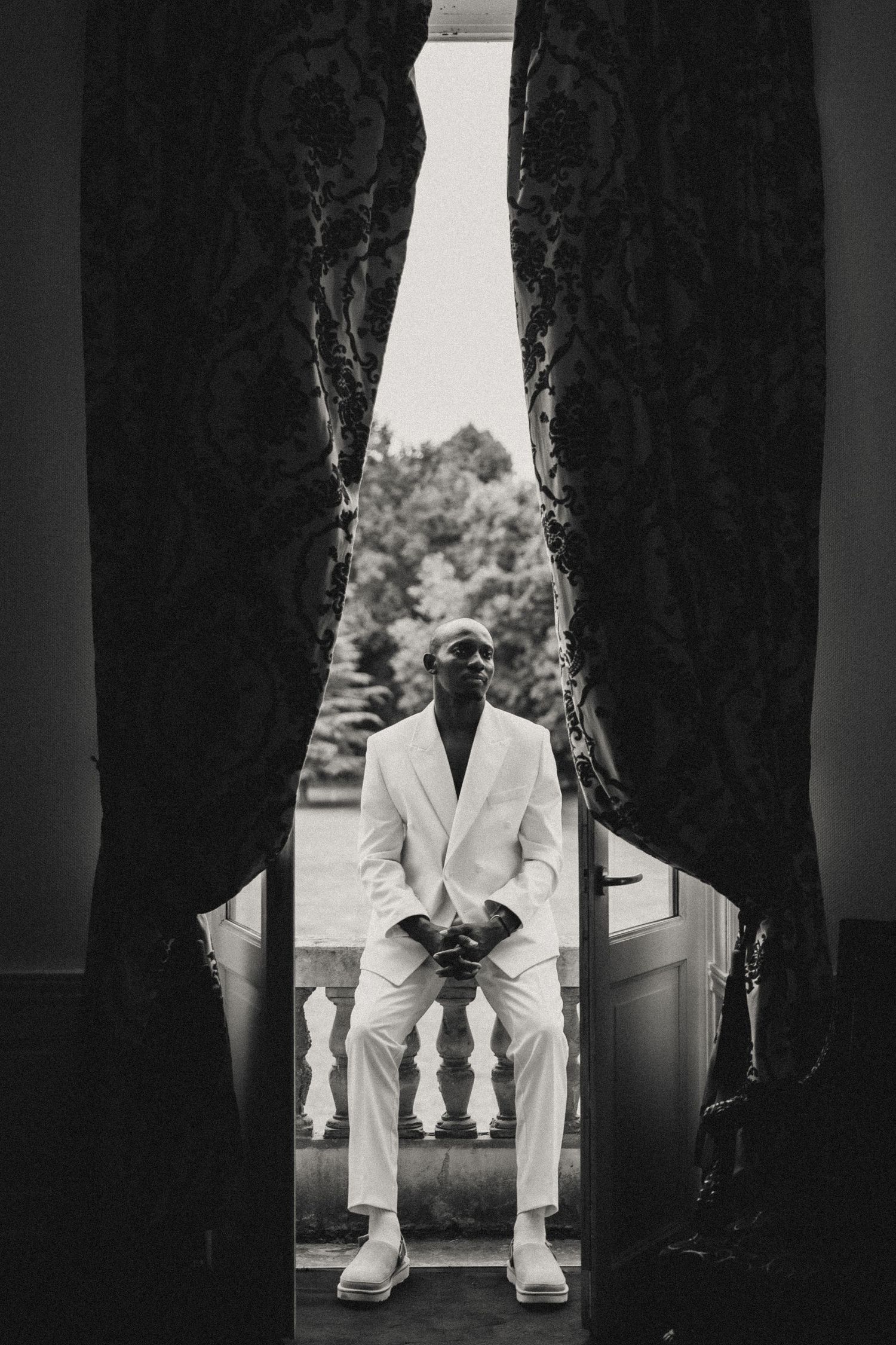 Black and white photo showing person in white suit seated between dark curtains.