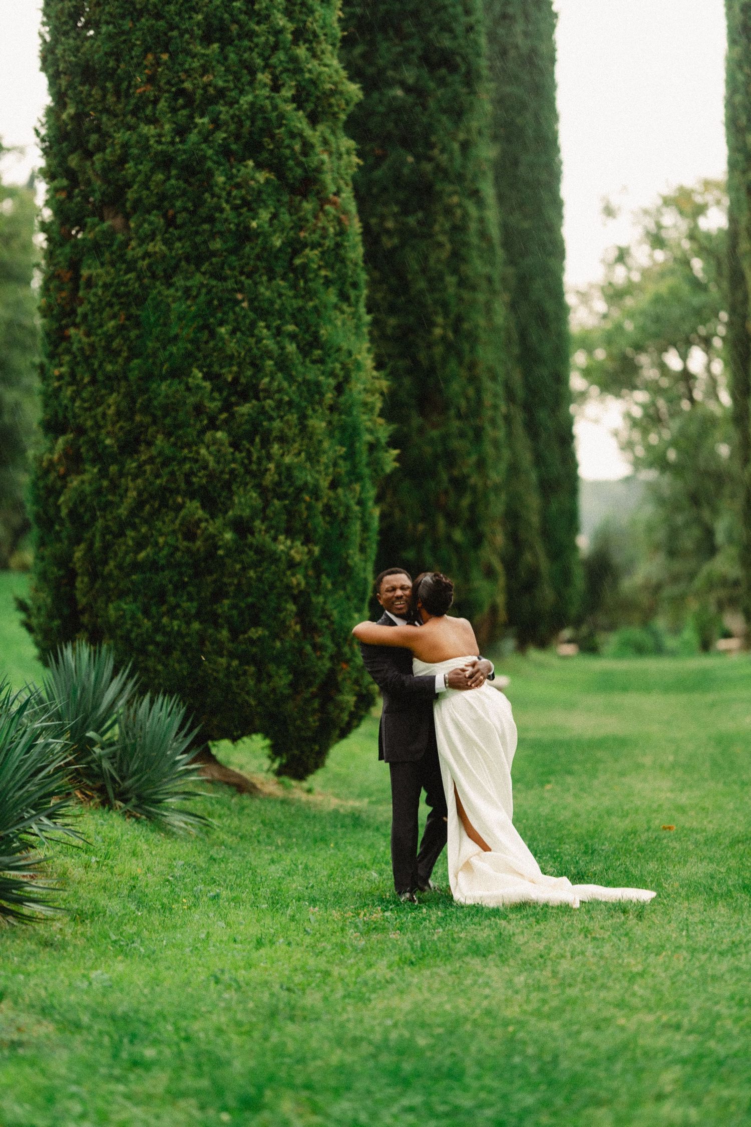 Wedding couple shares intimate moments while sitting on grass surrounded by towering cypress trees.