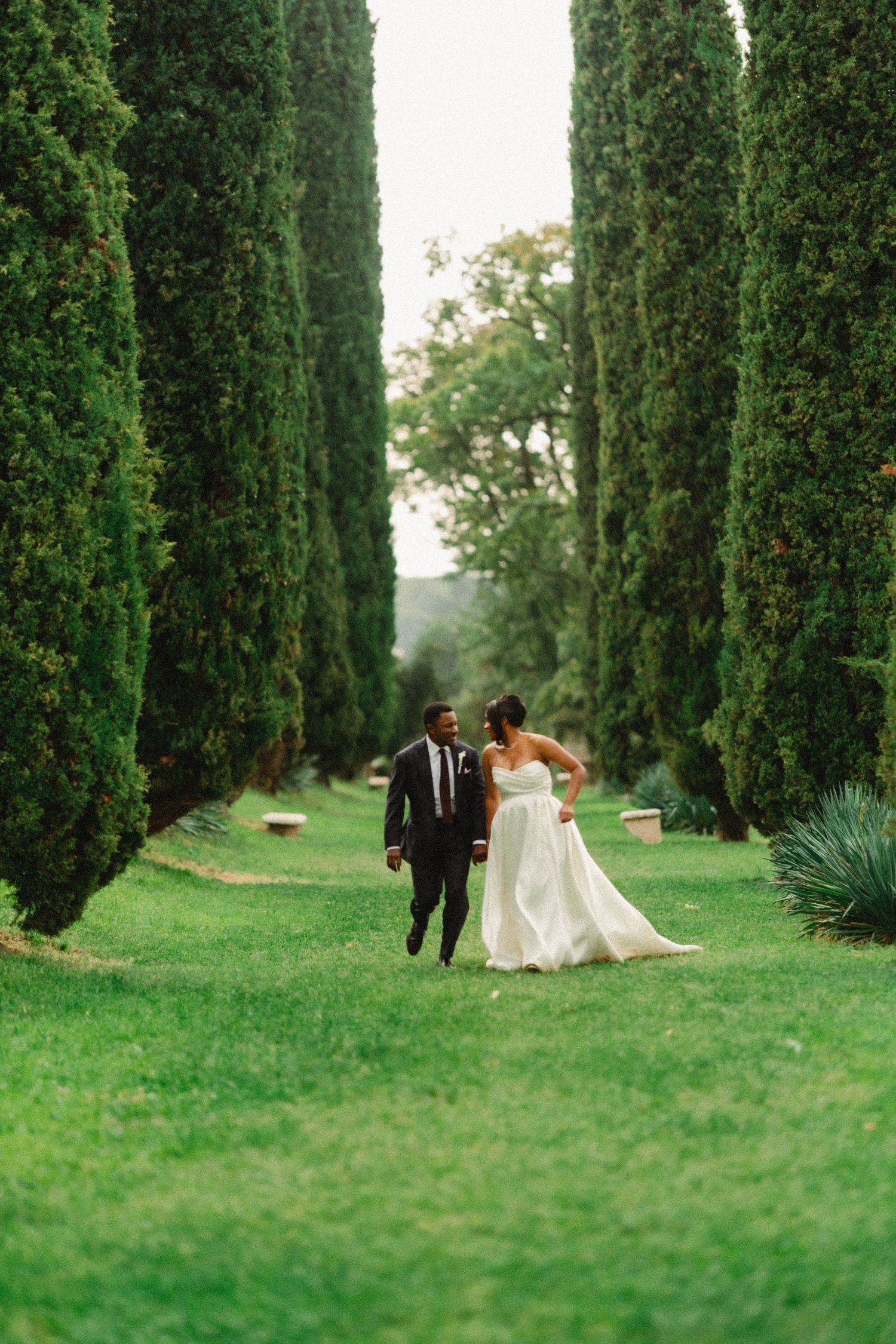 A romantic wedding photo sequence showing a couple walking and sitting among tall cypress trees on a lush green lawn.