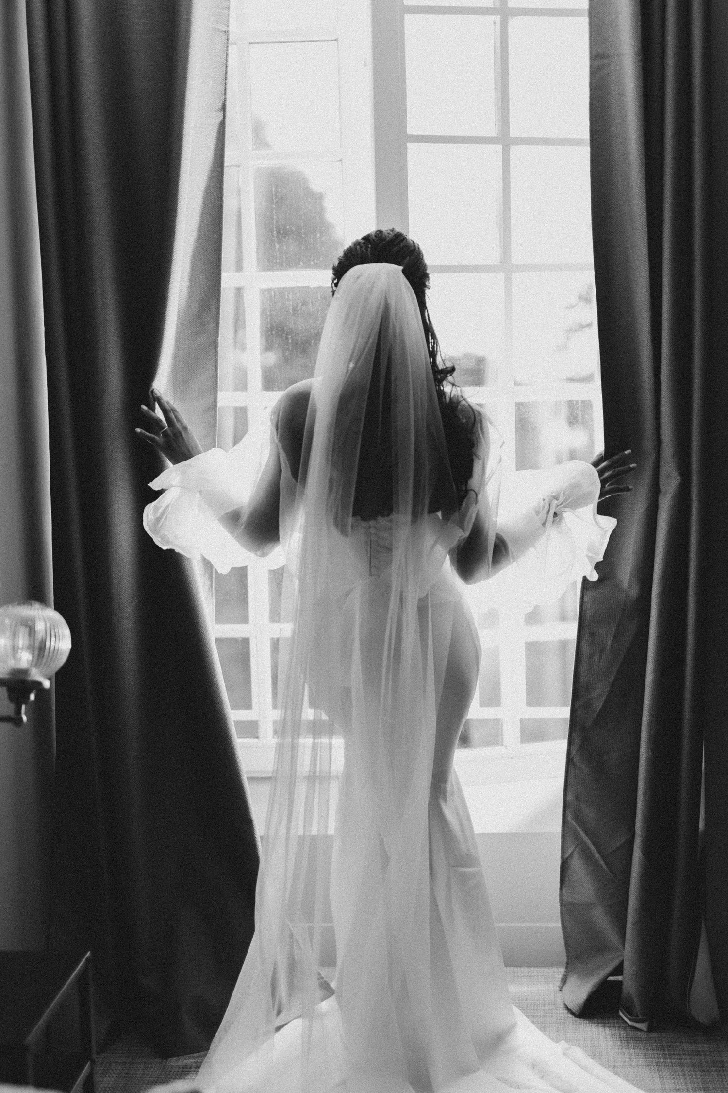 Silhouette of a bride in front of bright windows with flowing veil and dramatic curtains.