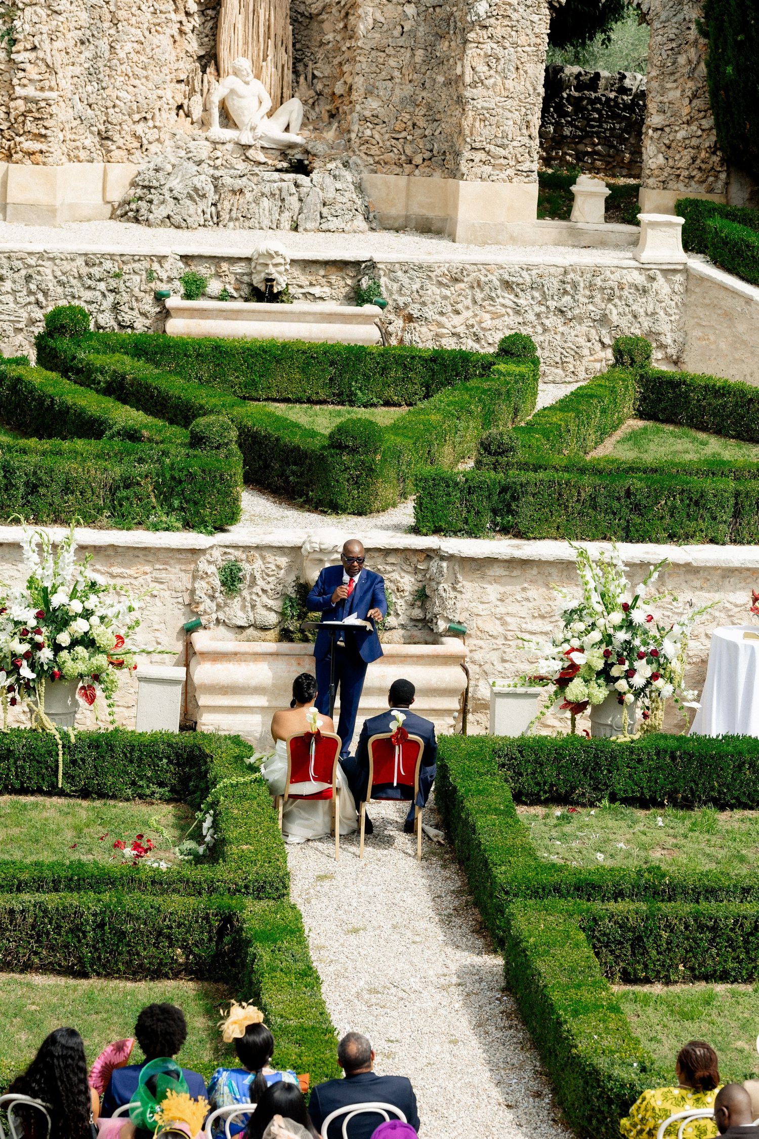 Outdoor garden wedding ceremony setup with stone walls, manicured hedges and white floral arrangements along gravel paths.
