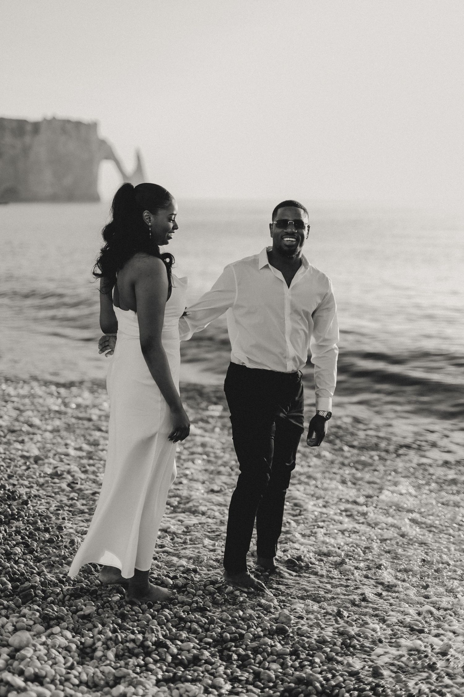 A couple in elegant attire poses on a beach with cliffs in the background.