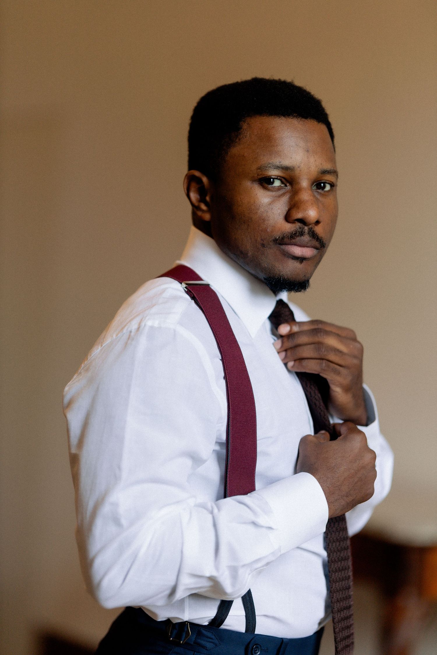 A person adjusts their burgundy tie while wearing a crisp white dress shirt and suspenders in a formal setting.