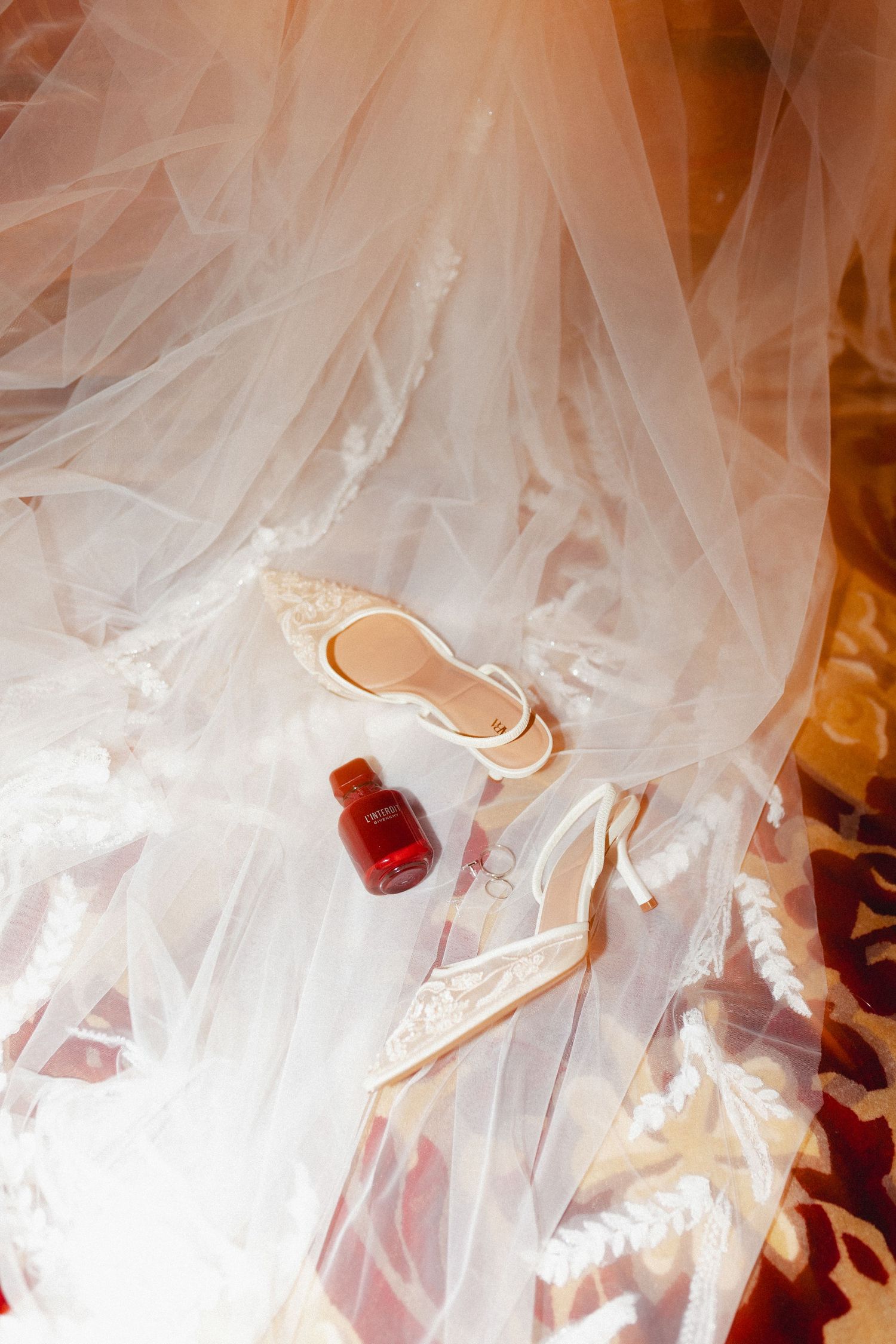 White wedding veil draped over elegant bridal shoes with a small red bottle on a patterned floor.