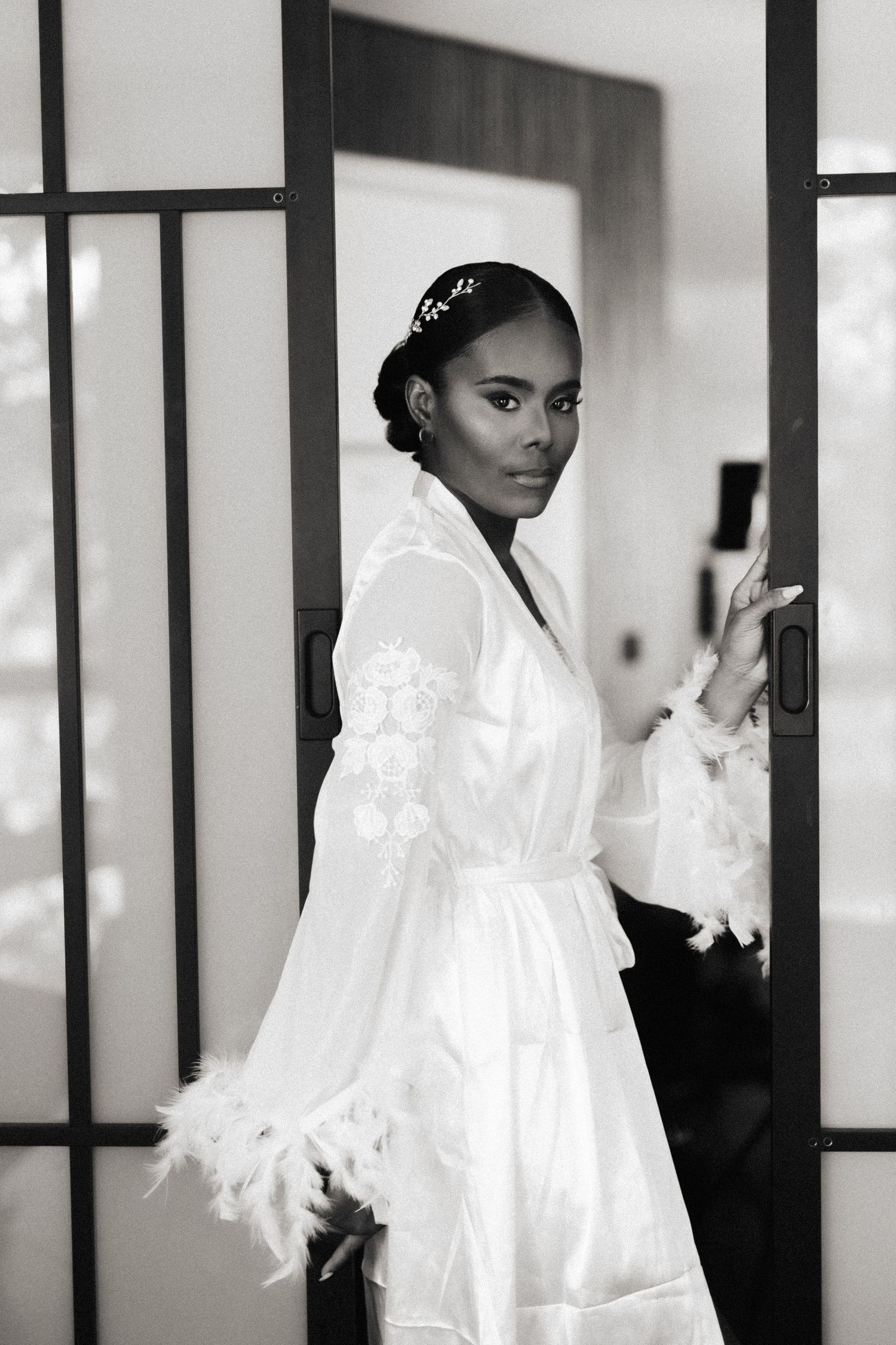 A black and white portrait of someone wearing an elegant white satin robe with feather details and a decorative headpiece.