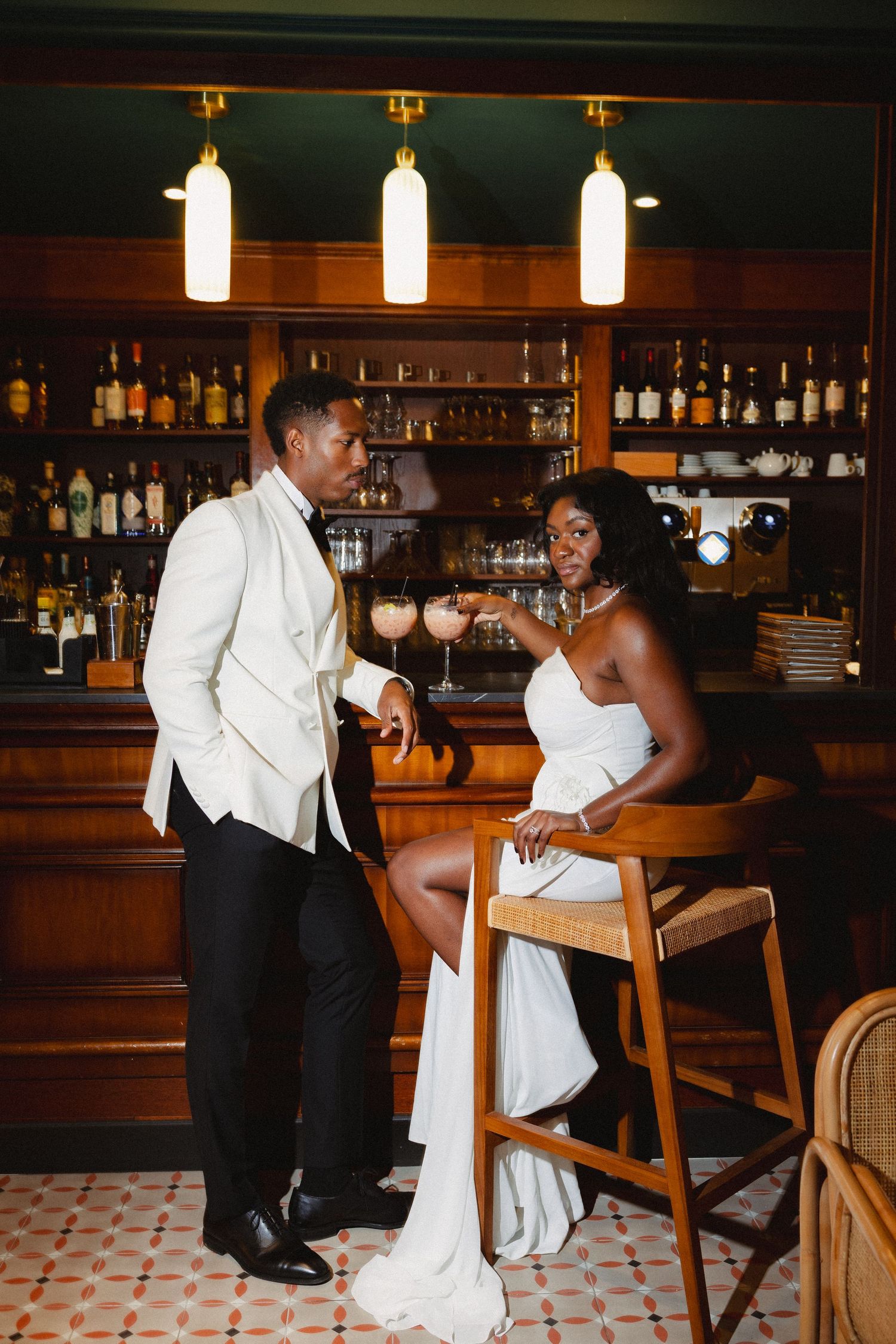 A couple in elegant white formal attire share a romantic moment at a stylish bar with pendant lighting and wooden decor.