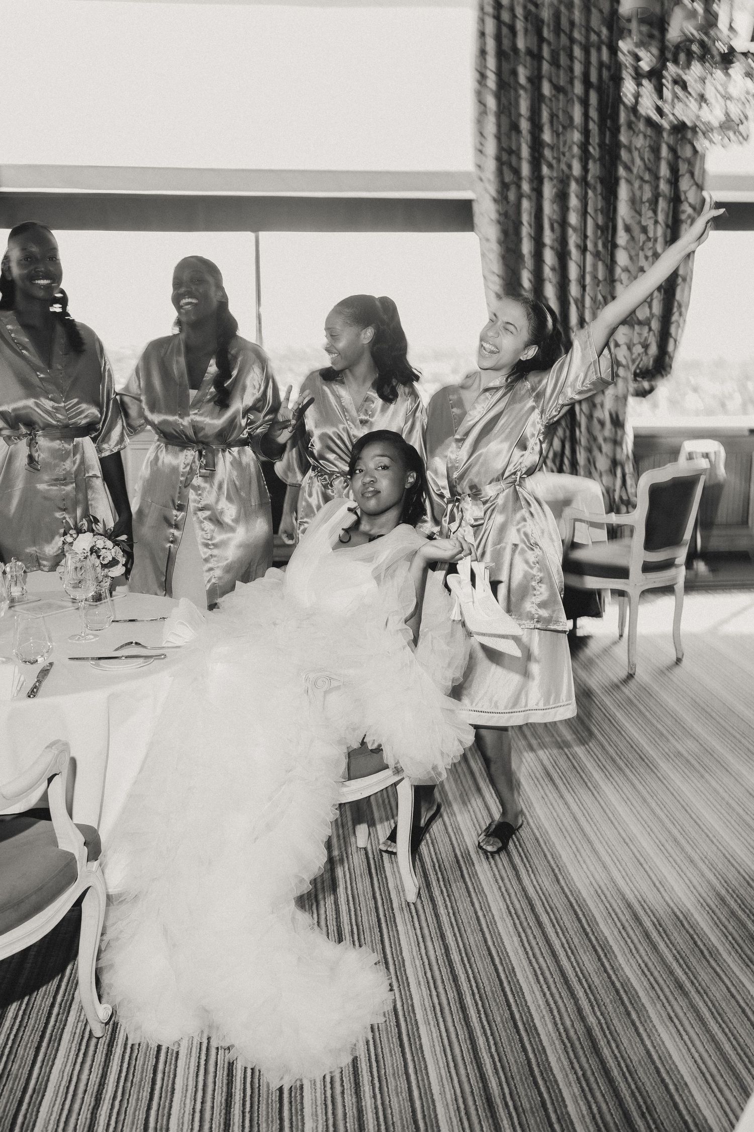 A joyful wedding celebration scene with bridesmaids in robes surrounding a bride in a voluminous white tulle dress.
