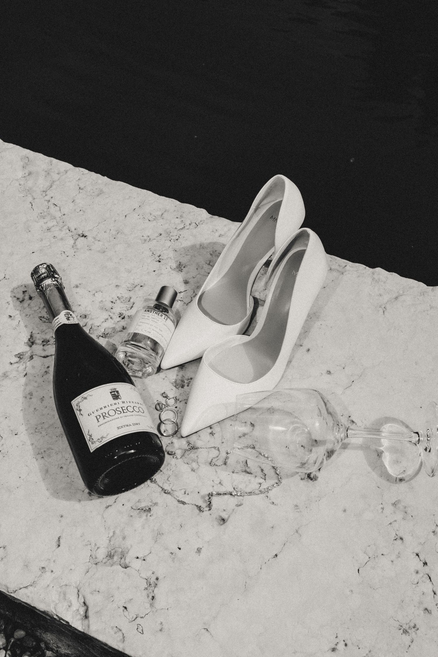 White stiletto heels arranged with champagne bottles on a textured floor in monochrome style.